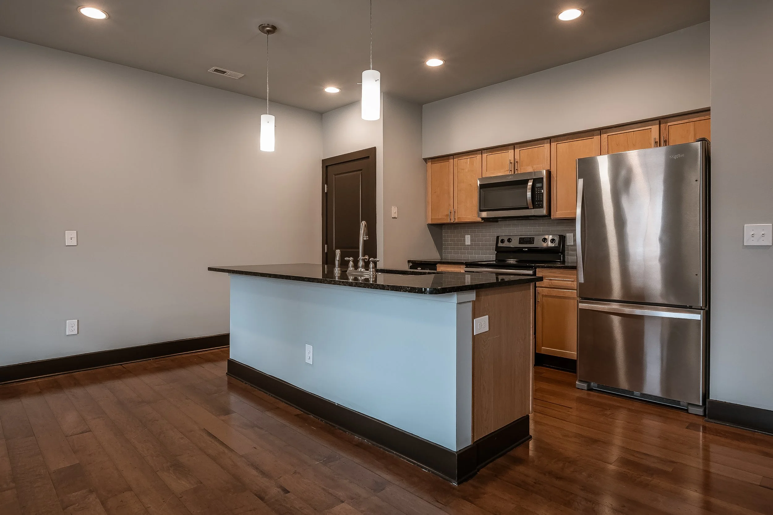 Modern kitchen with hardwood floors, gray walls, ceiling lights, centered island with black countertop, stainless steel refrigerator, microwave, and stove, wooden cabinets, and a gray tile backsplash.
