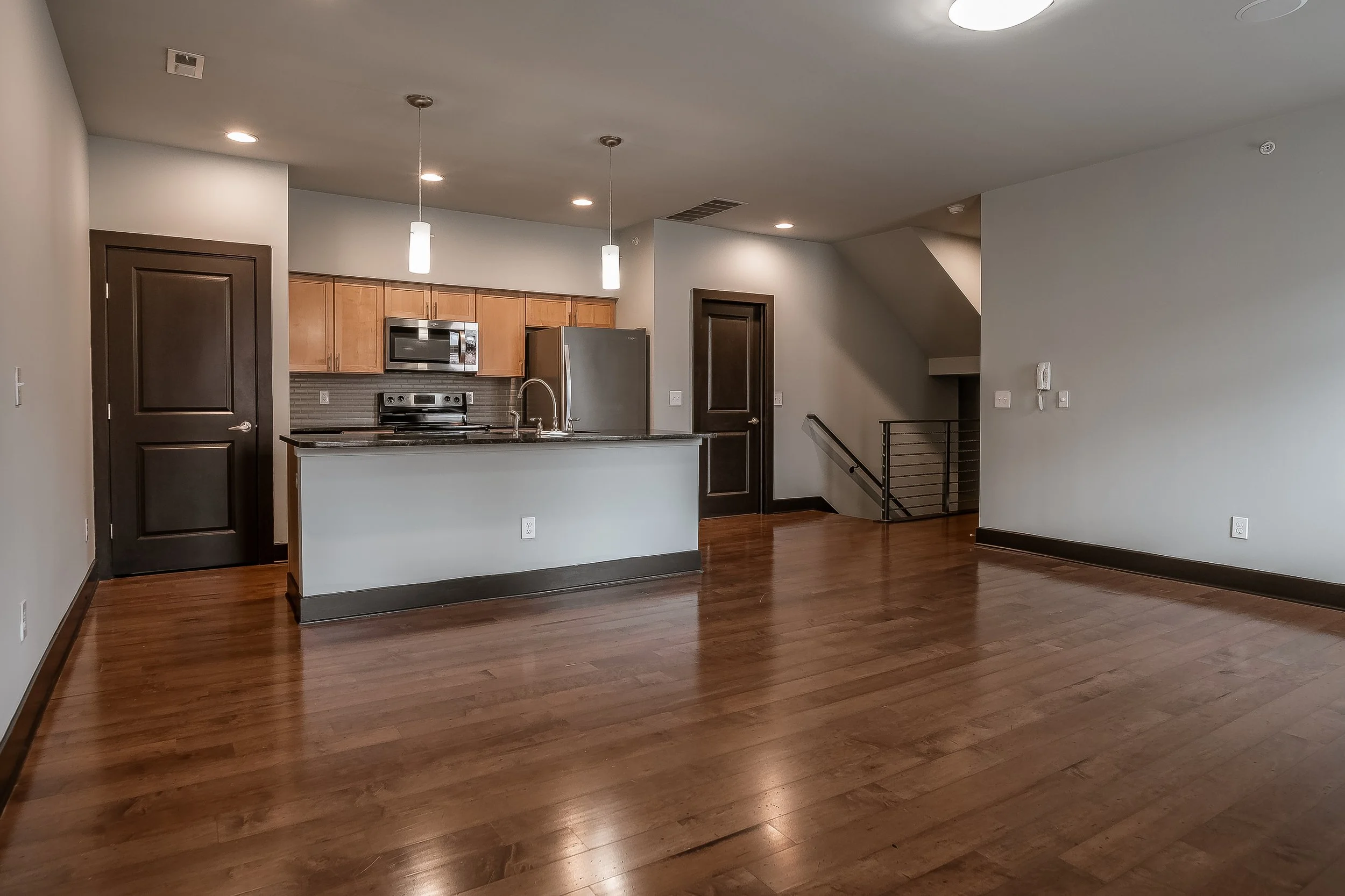 Empty living room and kitchen area with hardwood floors, modern dark wood doors, and kitchen cabinets, stainless steel appliances, and pendant lights.