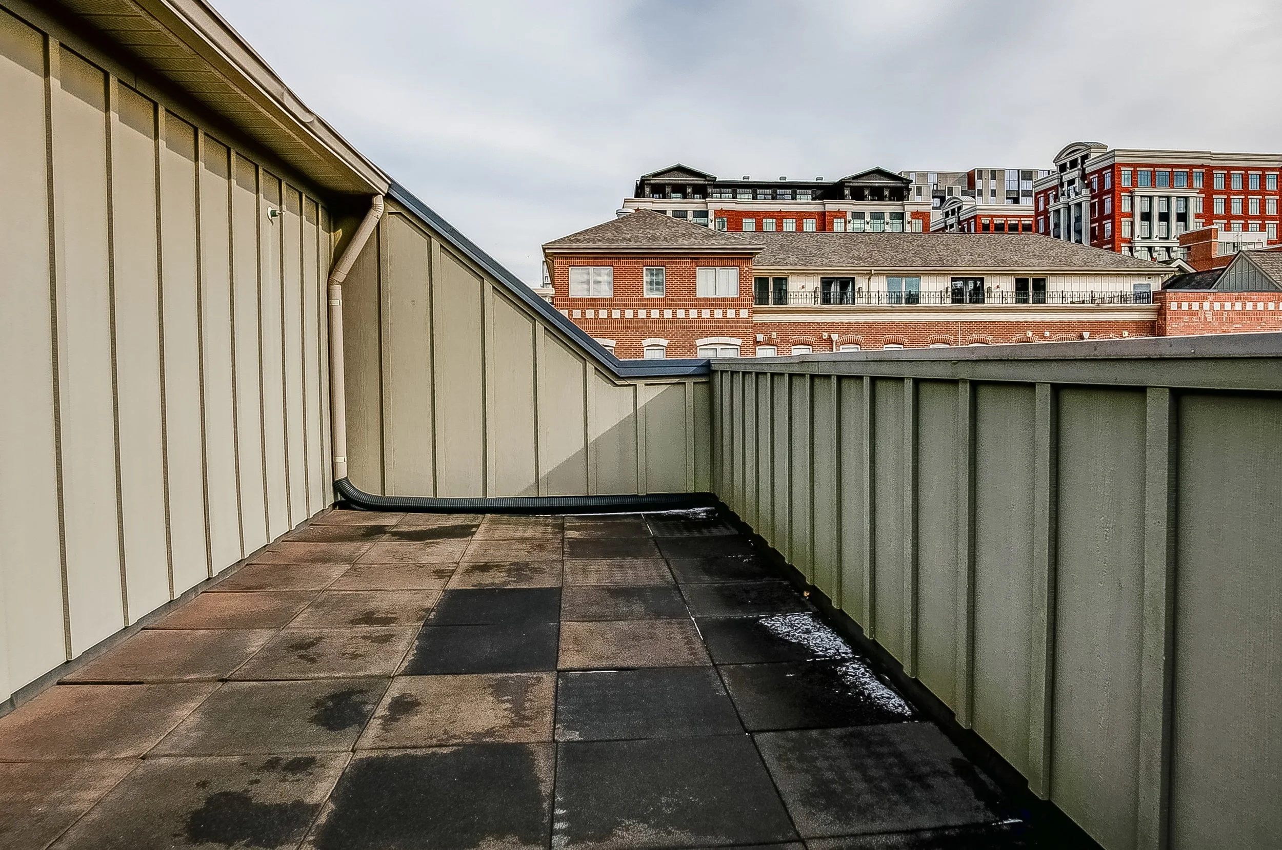 Rooftop balcony with beige siding, black drainage pipe, and tiled flooring, overlooking neighboring buildings with brick and modern architecture.