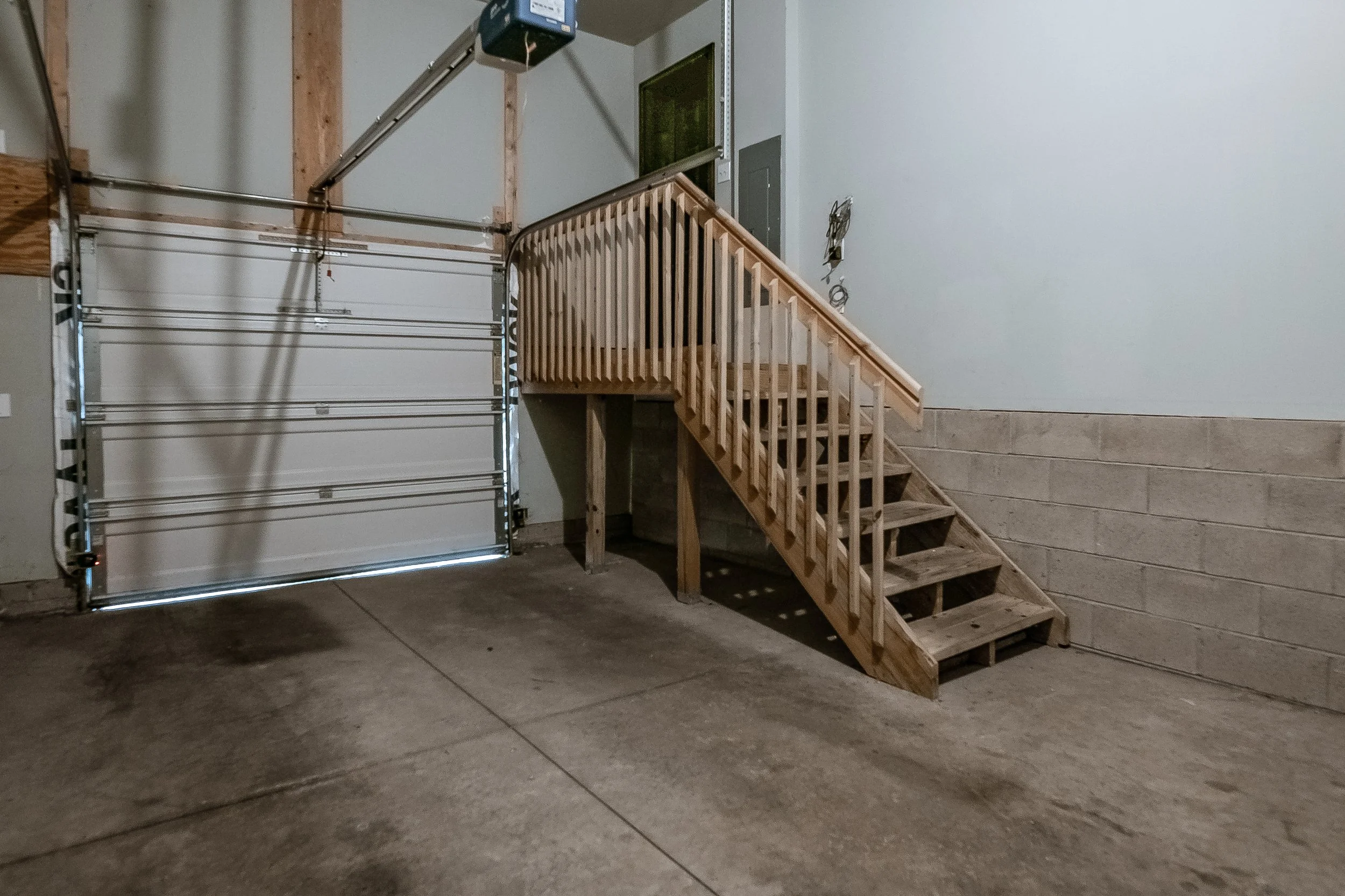 Empty garage with a metal roll-up door, concrete floor with dark stains, and a wooden staircase leading to an upper level, with a gray wall on the right side.