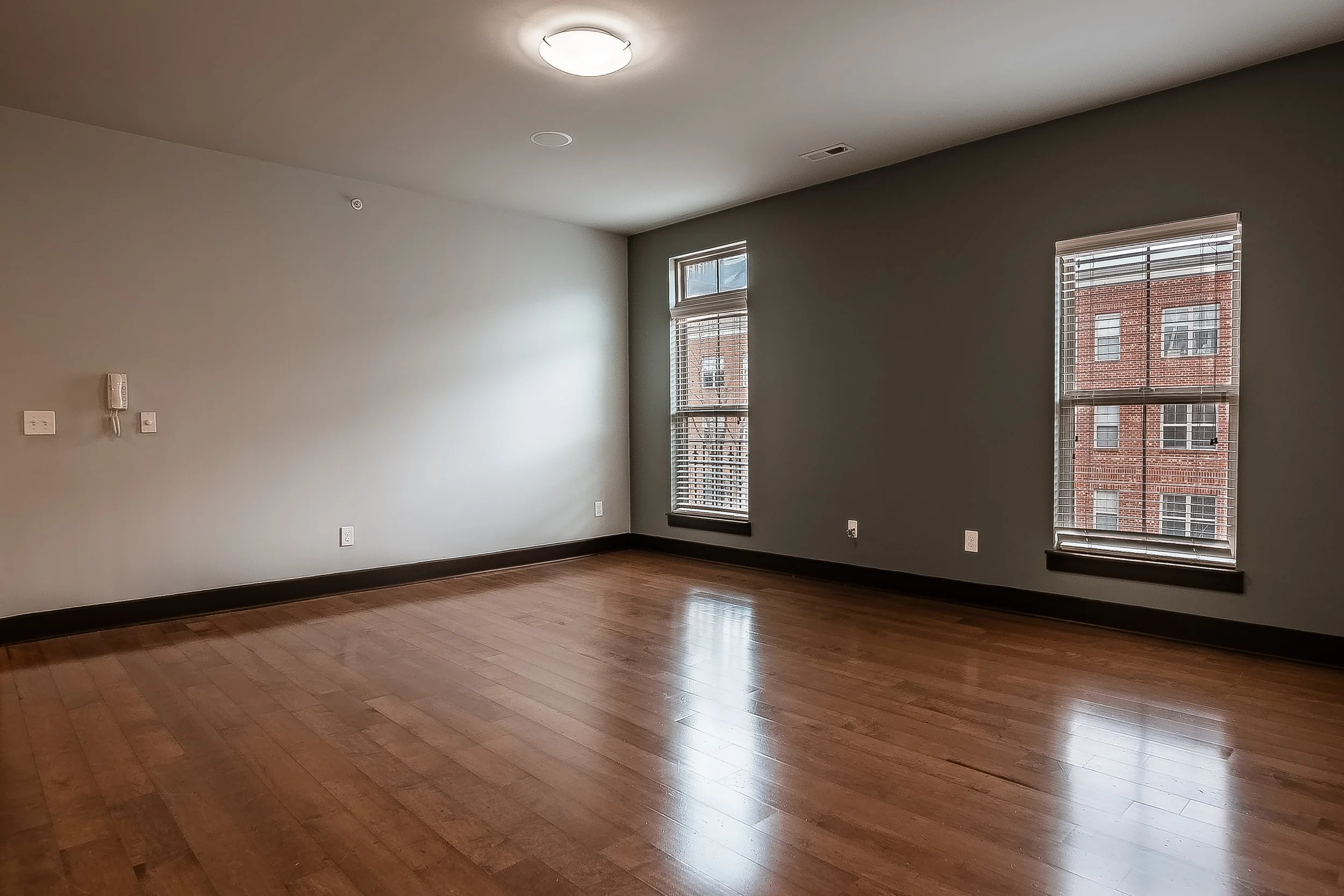 Empty living room with hardwood floors, concrete walls, two large windows with blinds, ceiling light, and some electrical outlets.