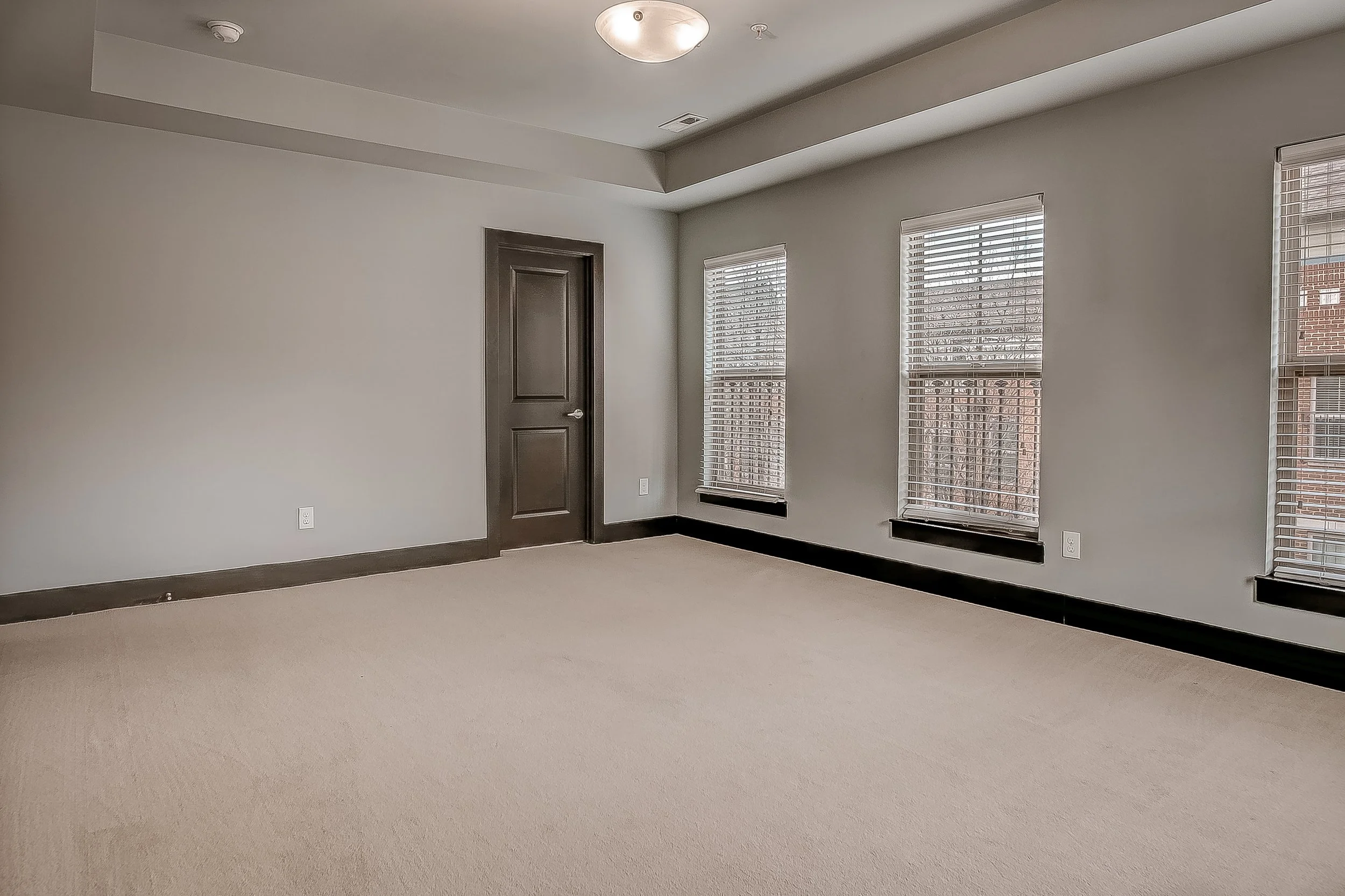 Empty room with beige carpet, light gray walls, three large windows with blinds, a dark brown door, and black baseboards, with natural light coming through the windows.