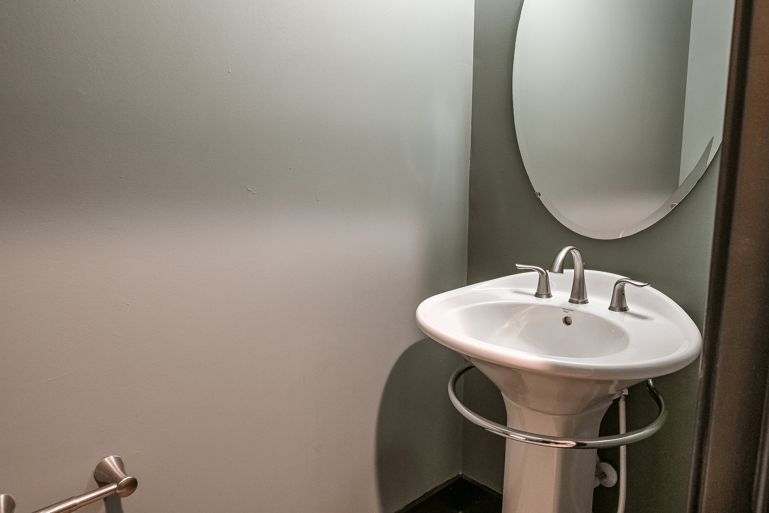 A white pedestal sink with chrome faucet and handles, a large oval mirror above it, and a towel bar to the left, in a bathroom with gray walls.