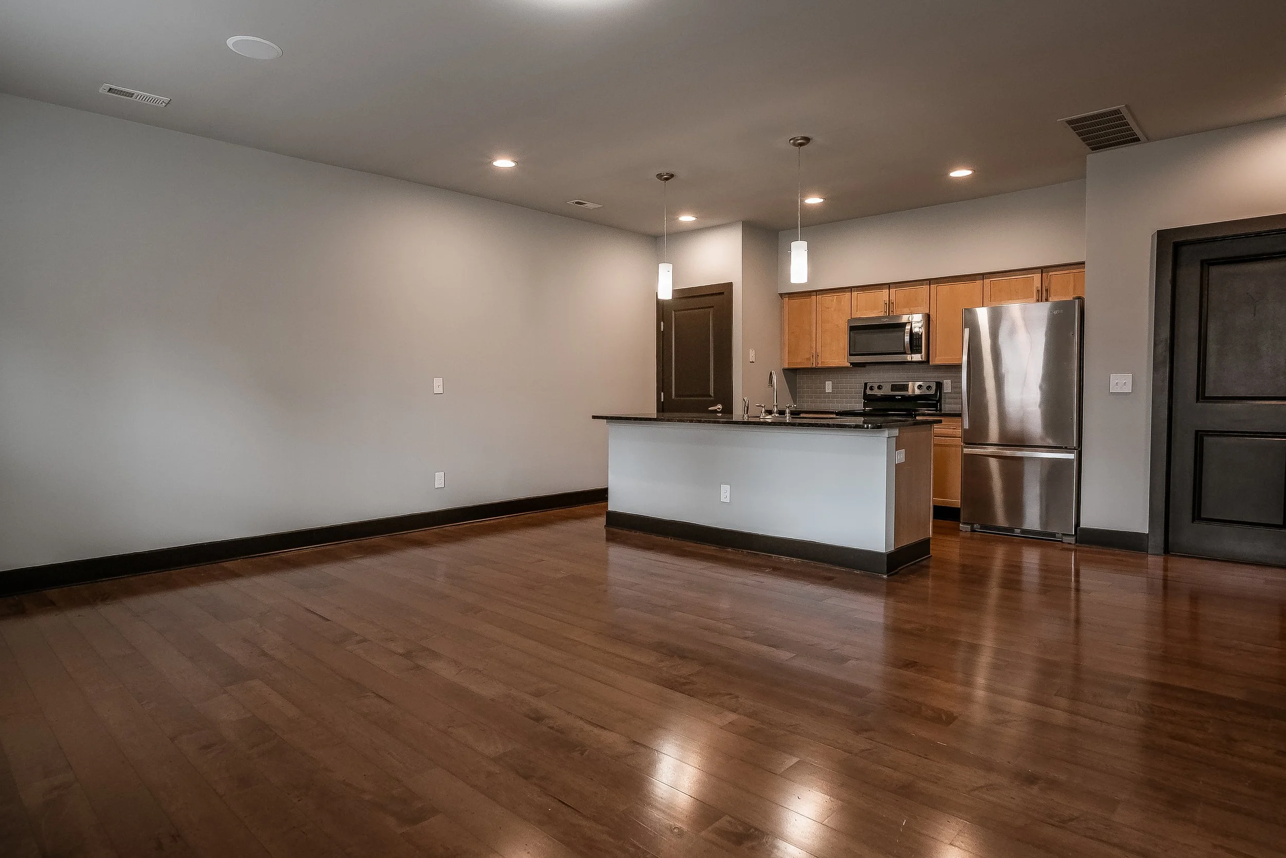 Empty living room with hardwood floors, kitchen with stainless steel appliances, wooden cabinets, and pendant lights.