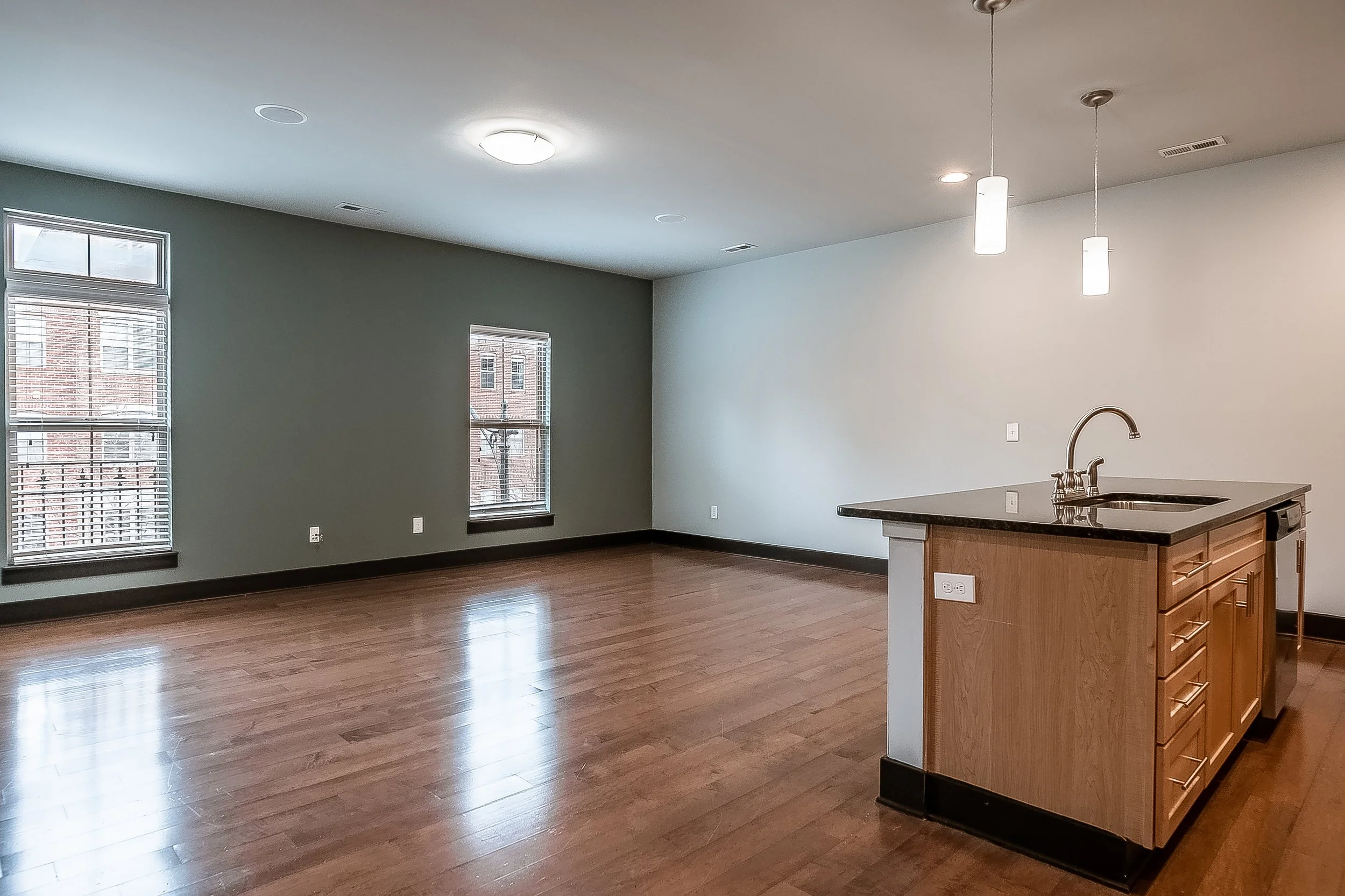Empty living room and kitchen area with hardwood floors, green and white walls, and windows providing natural light. The kitchen has a small island with a sink and pendant lighting.