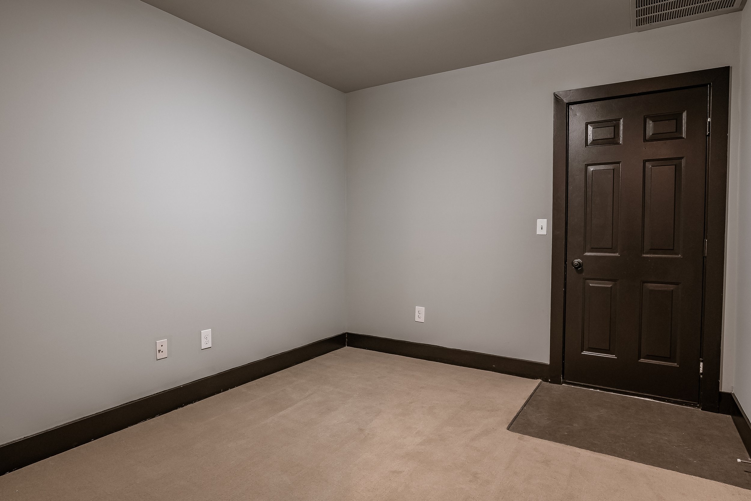 Empty room with beige carpet, white walls, dark brown door, and baseboards, with electrical outlets and a vent on the ceiling.