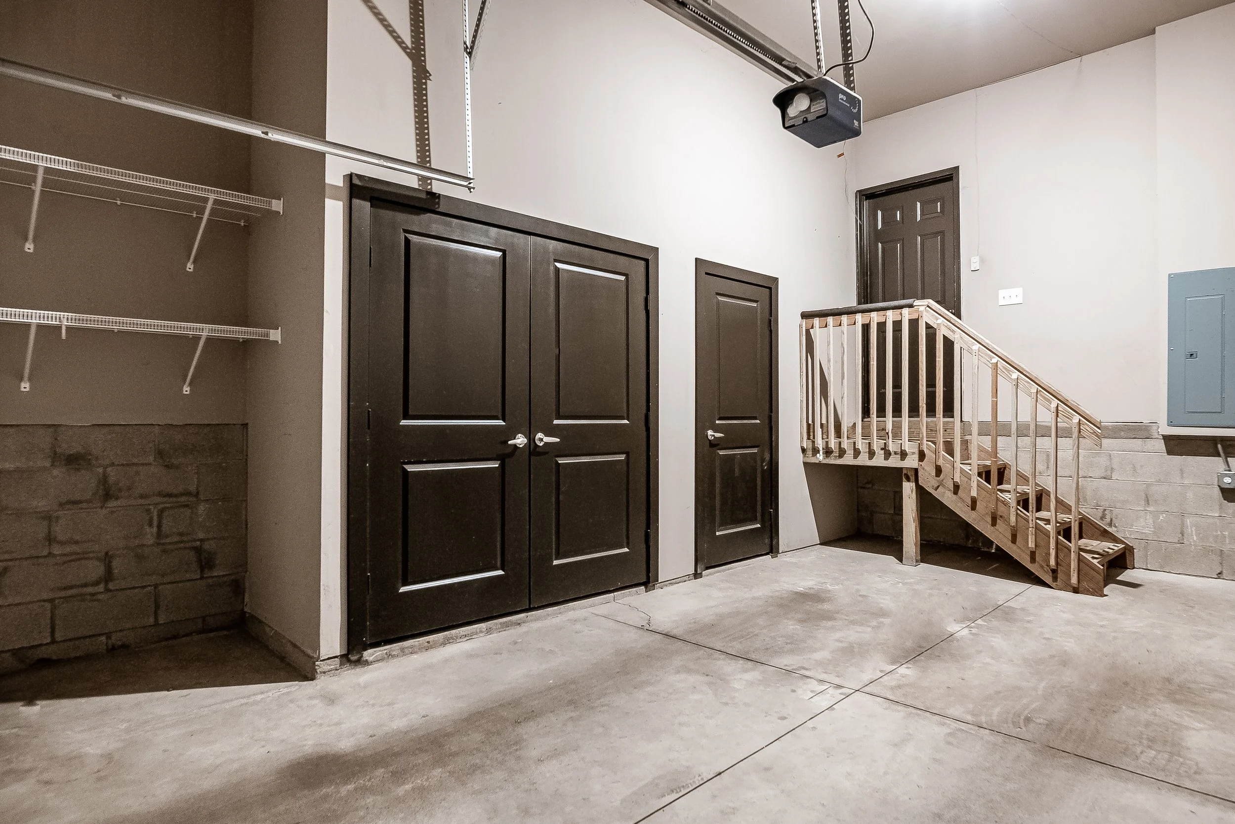 Interior of a garage with black double doors, a smaller black door, a wooden staircase leading to a door, a wire shelf on the left, an electric garage door opener on the ceiling, and a gray electrical box on the wall.