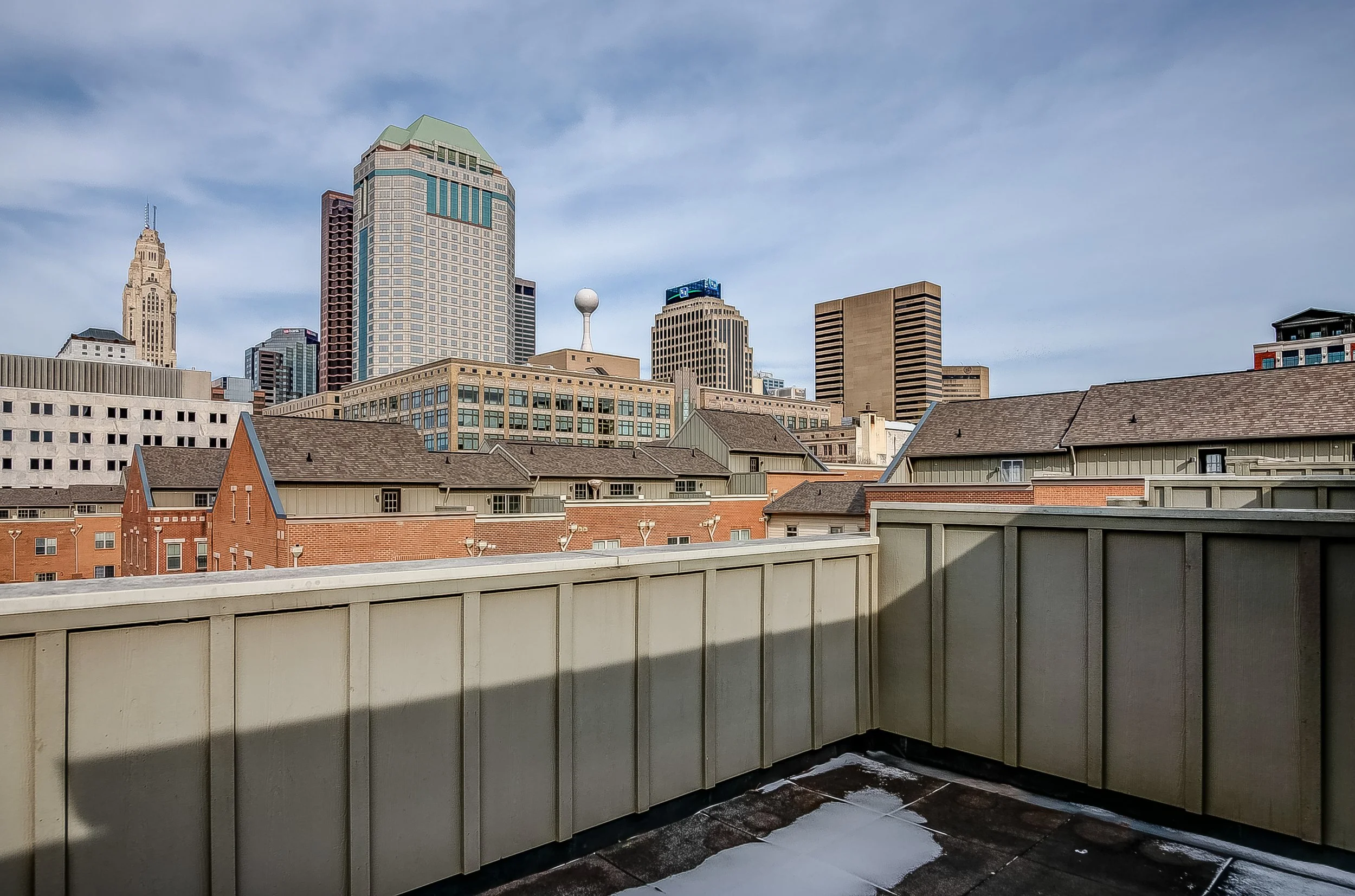 City skyline with tall modern buildings seen from a rooftop balcony with railings and some patches of snow on the ground.