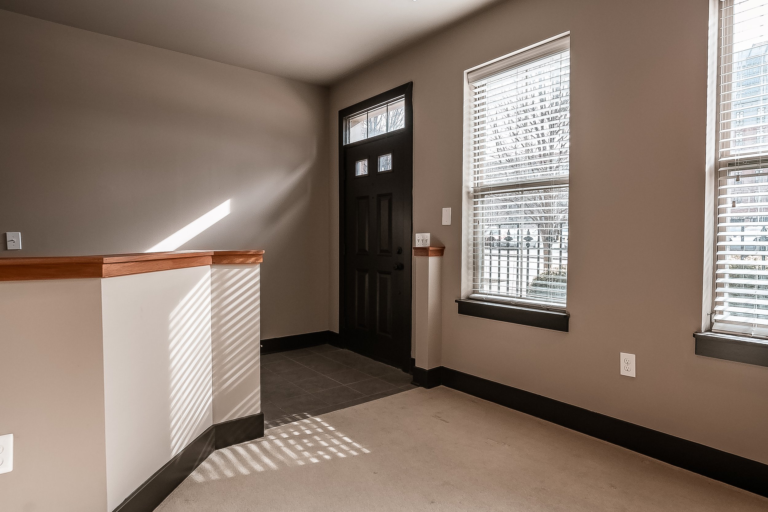 Empty room with two windows, a black front door, and a corner with a half wall, beige carpet, and dark trim.