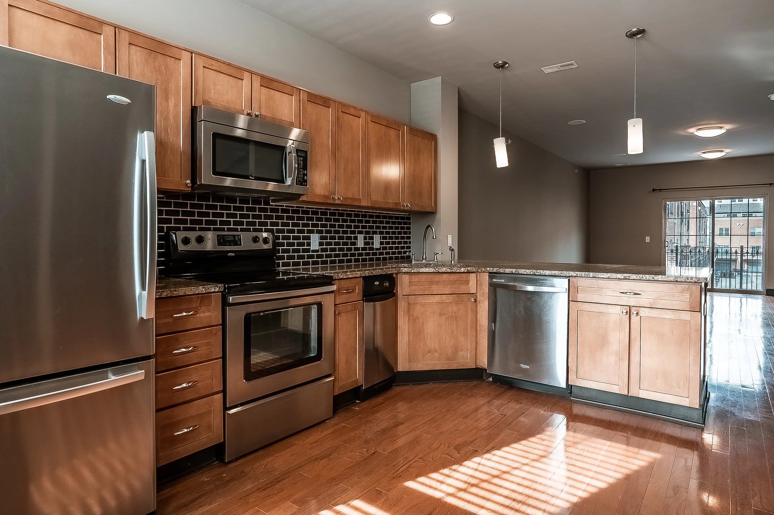 Modern kitchen with wooden cabinets, stainless steel appliances, granite countertops, and hardwood floors, illuminated by hanging pendant lights and natural light from a sliding glass door.