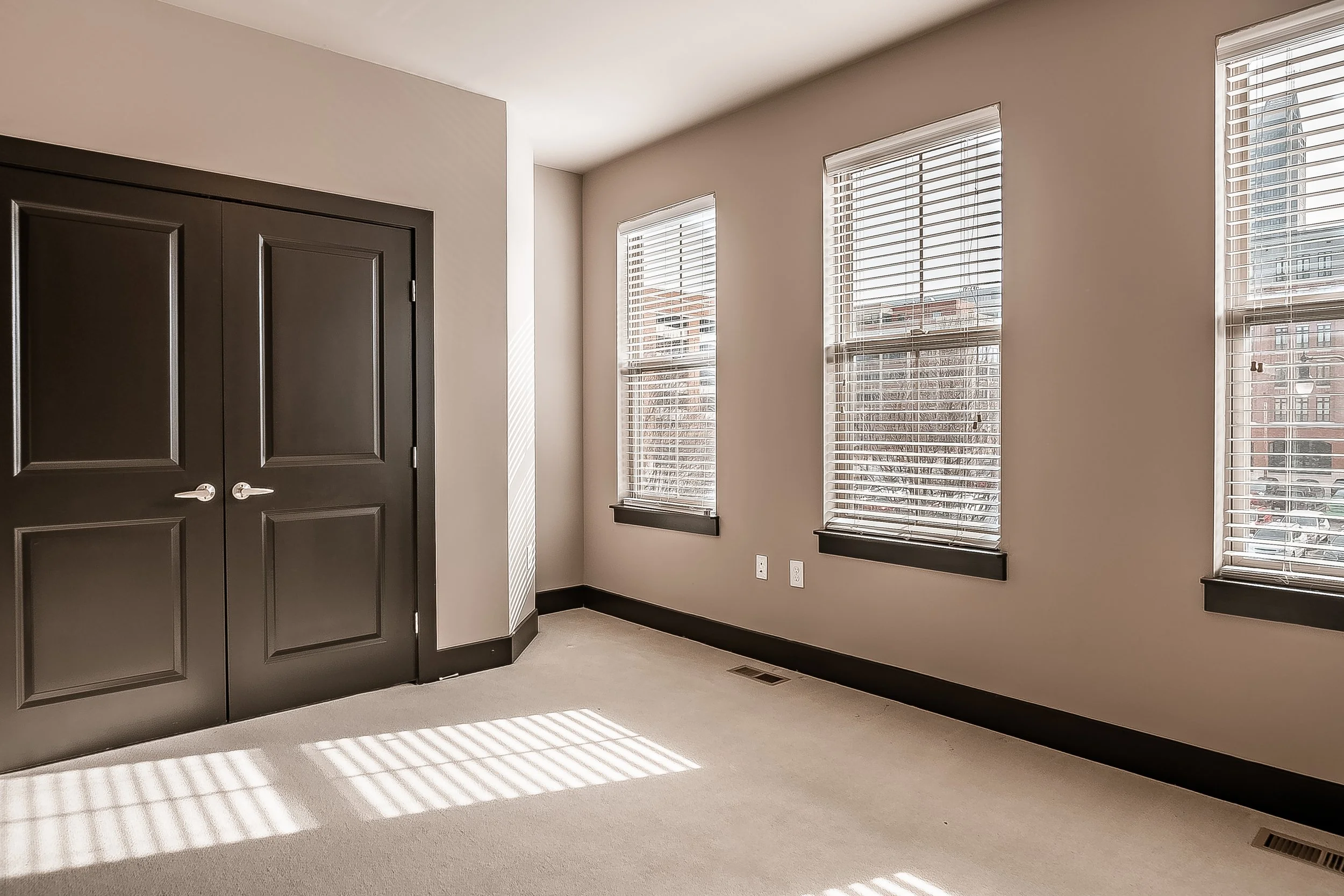 Empty room with beige carpet, three large windows with blinds, and dark brown door with double panels.