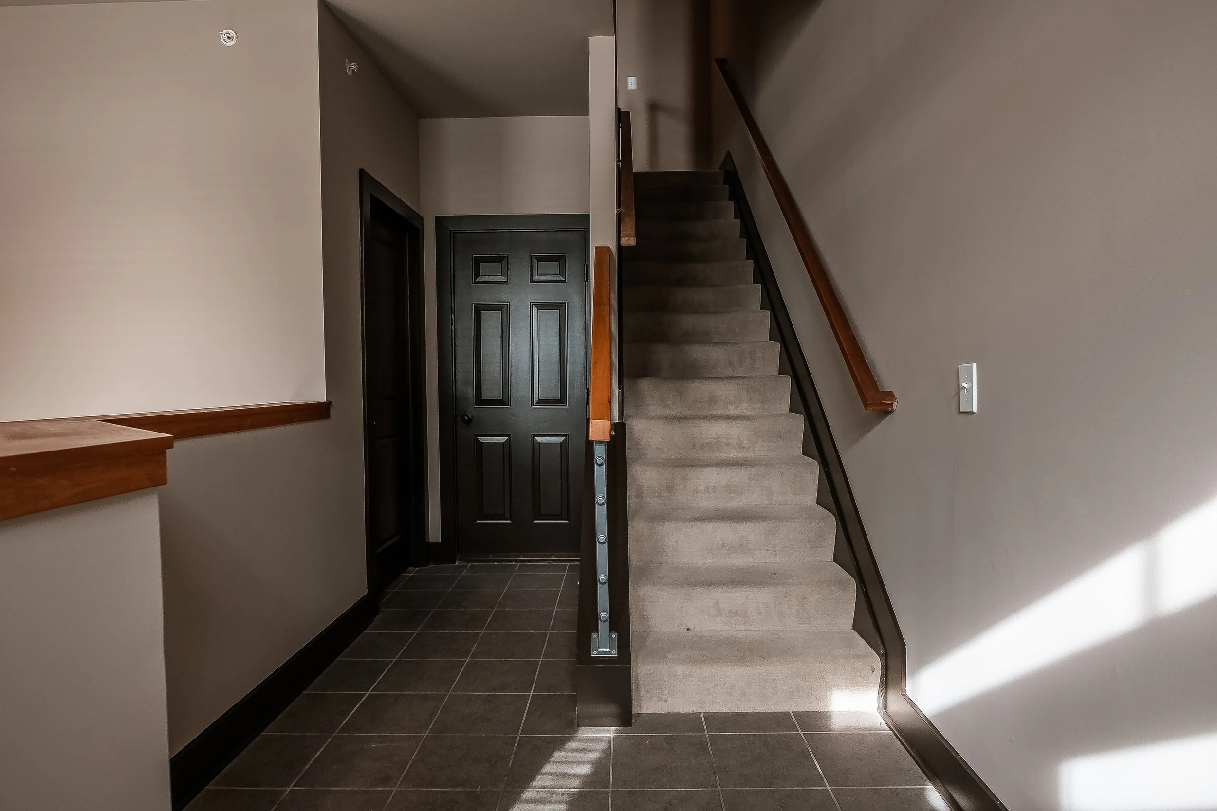 Interior view of a residential staircase with beige carpet, dark wood and black metal banister, black door at the base, and tiled floor.