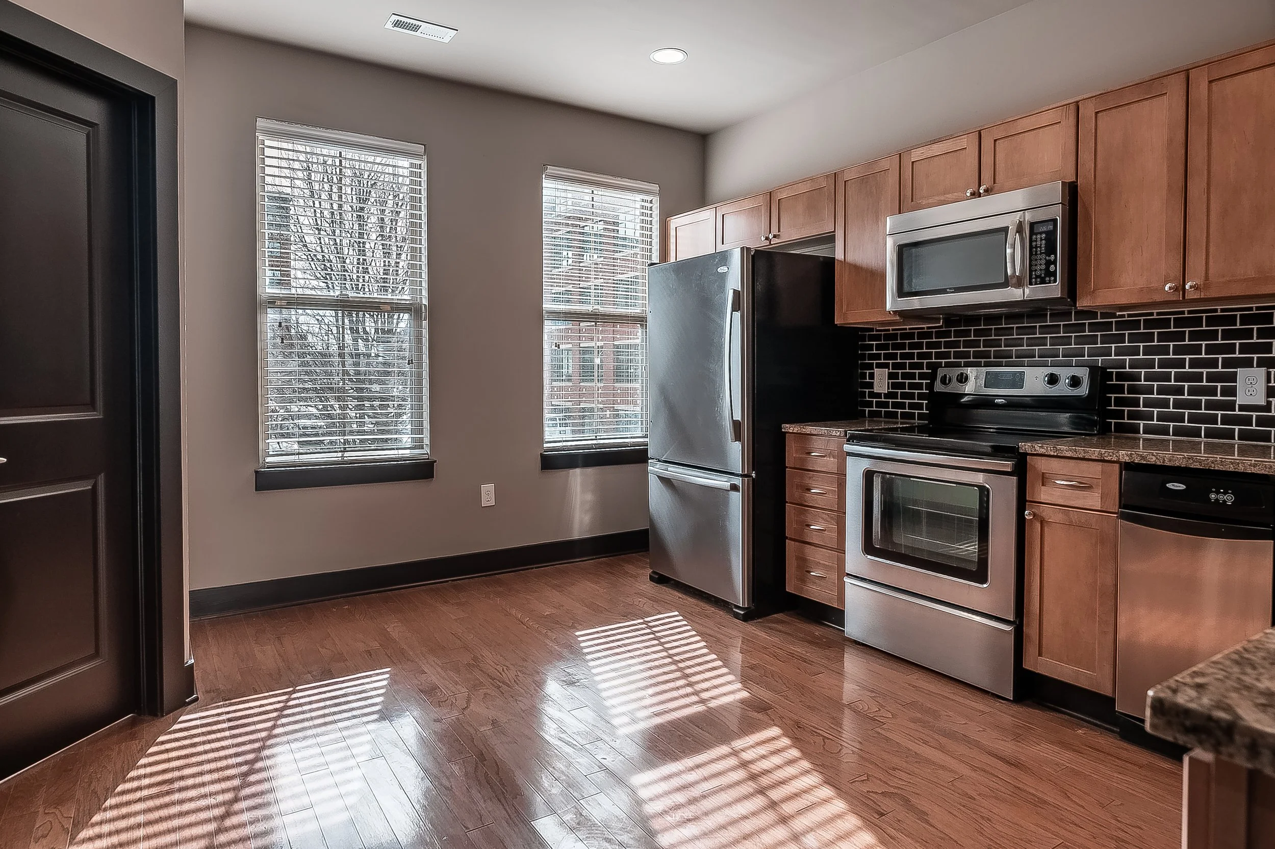 Kitchen with wooden cabinets, stainless steel refrigerator, microwave, stove, black tiled backsplash, hardwood floor, and two windows with blinds.