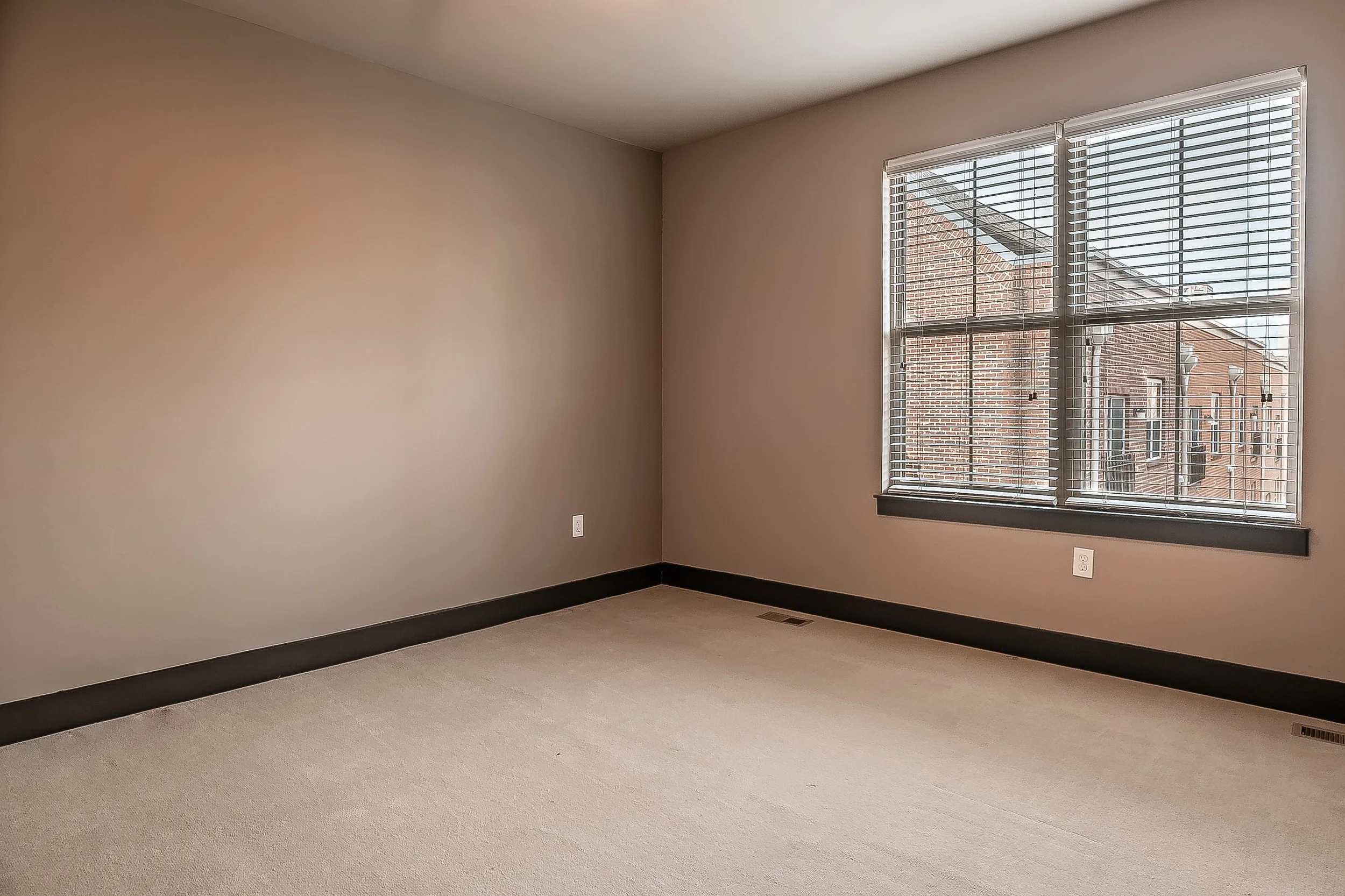 Empty room with beige carpet, beige walls, black trim, large window with blinds, view of brick building outside.