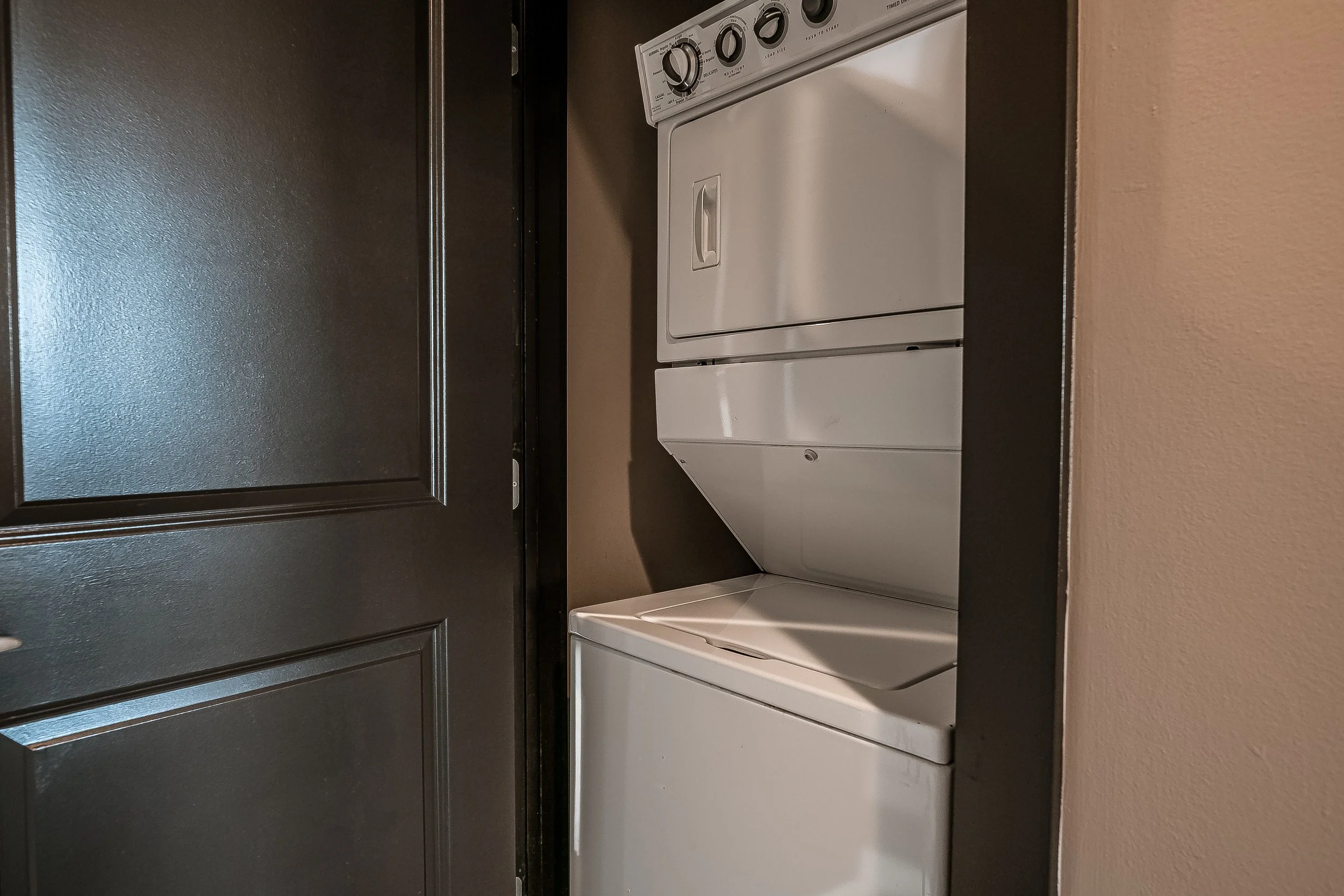 A stacked washer and dryer unit in a small laundry closet with a black door and beige wall.