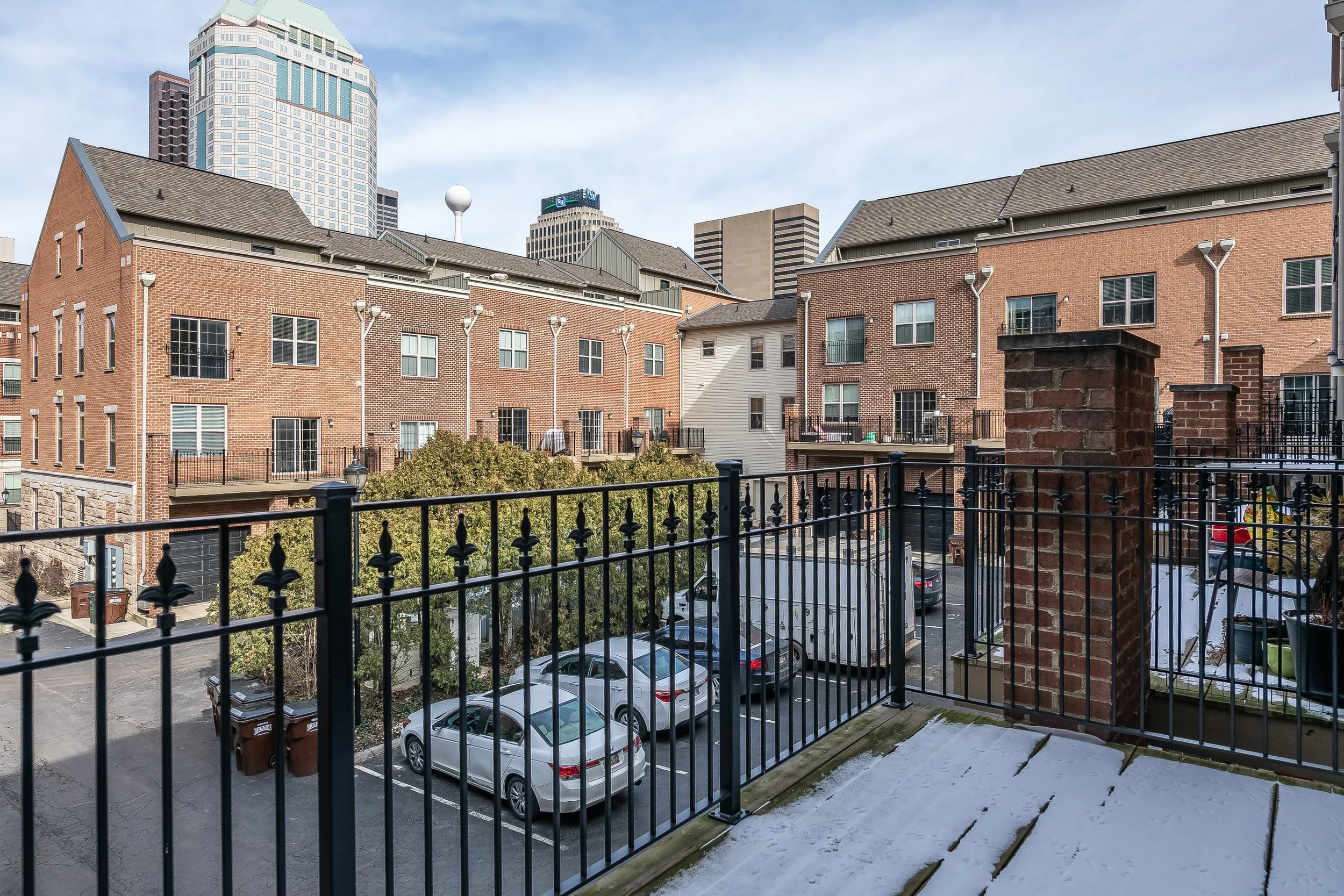 View from a balcony with a black metal railing showing a parking lot with cars, brick and white residential buildings, trees, and a city skyline with tall buildings in the background under a partly cloudy sky.