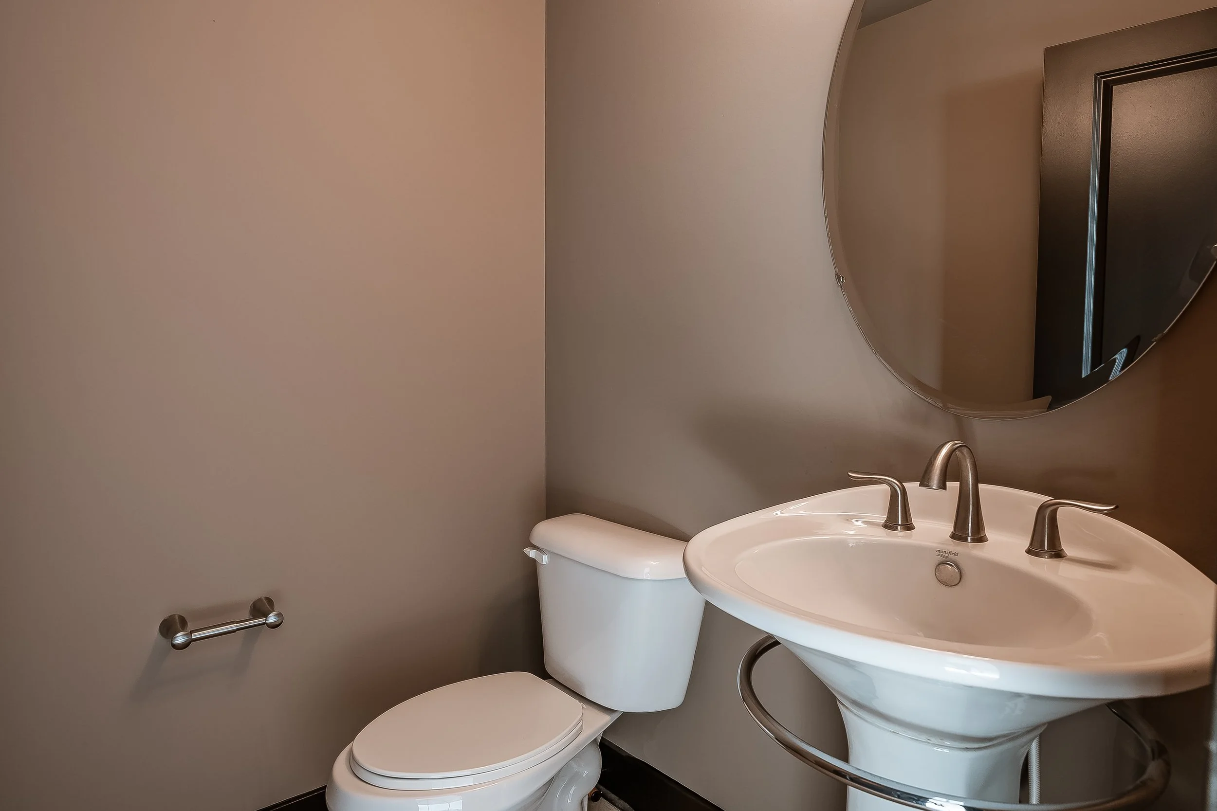 Bathroom with a white toilet, a white pedestal sink with three silver faucets, and a large round mirror on a beige wall.