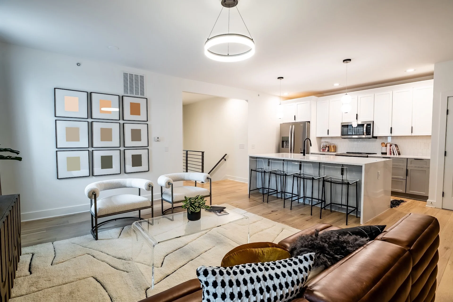 Modern living room with a tan leather sofa, white armchairs, a glass coffee table, and a large geometric rug. Art on the wall, potted plant, and an open kitchen with white cabinets, island with barstools, stainless steel appliances, and pendant lighting.