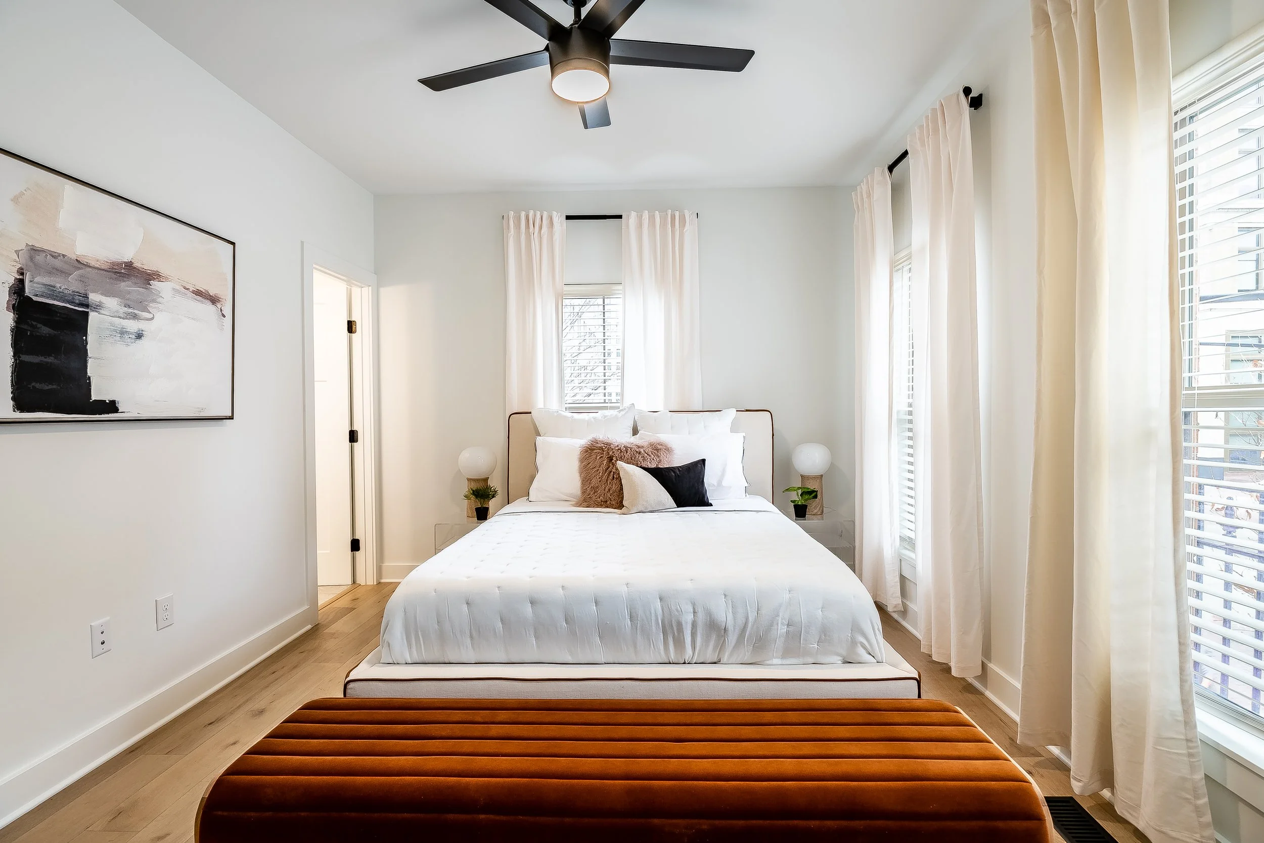 Bright bedroom with a bed featuring white bedding, pillows, and a brown faux fur pillow, flanked by two nightstands with white lamps and small green plants, beige curtains, hardwood floors, wall art, and a black ceiling fan.
