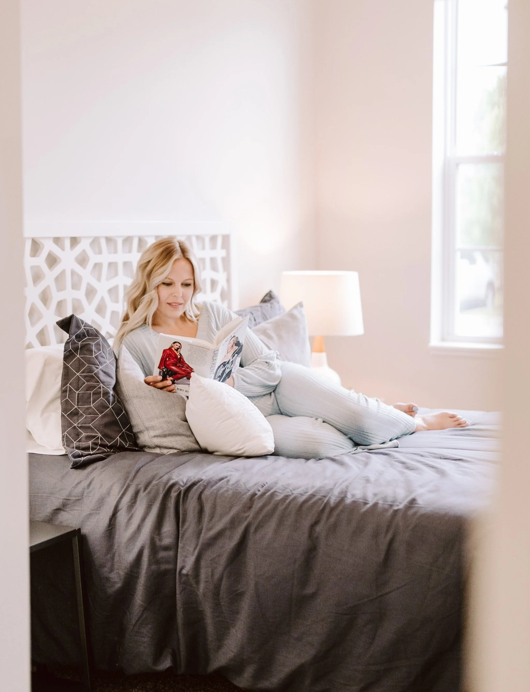 A woman with blonde hair lying on her side on a bed, reading a magazine in a well-lit bedroom with a window, nightstand, and lamp.