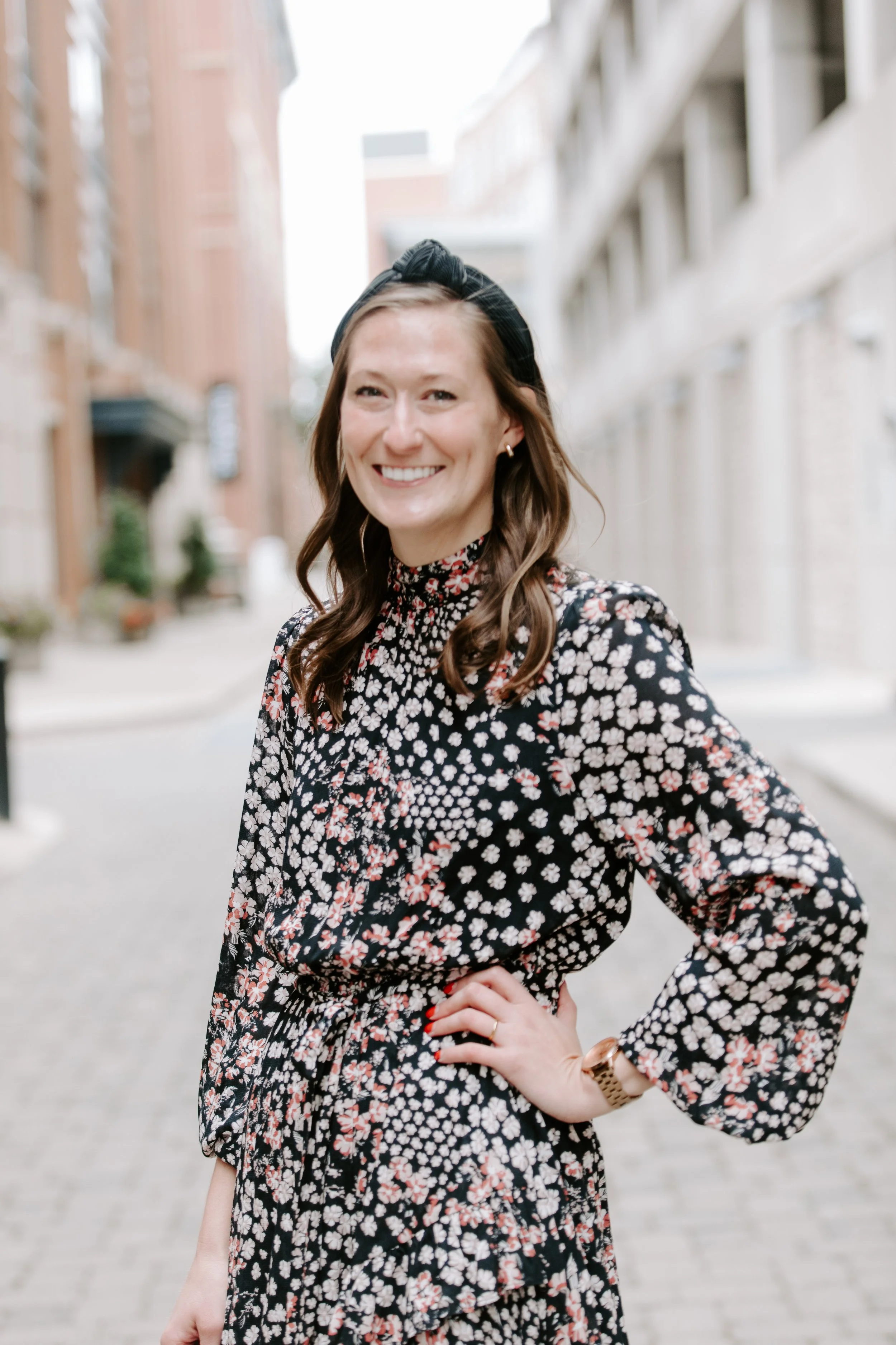 A woman smiling outdoors in an urban setting with brick buildings, wearing a floral dress and a black headband.