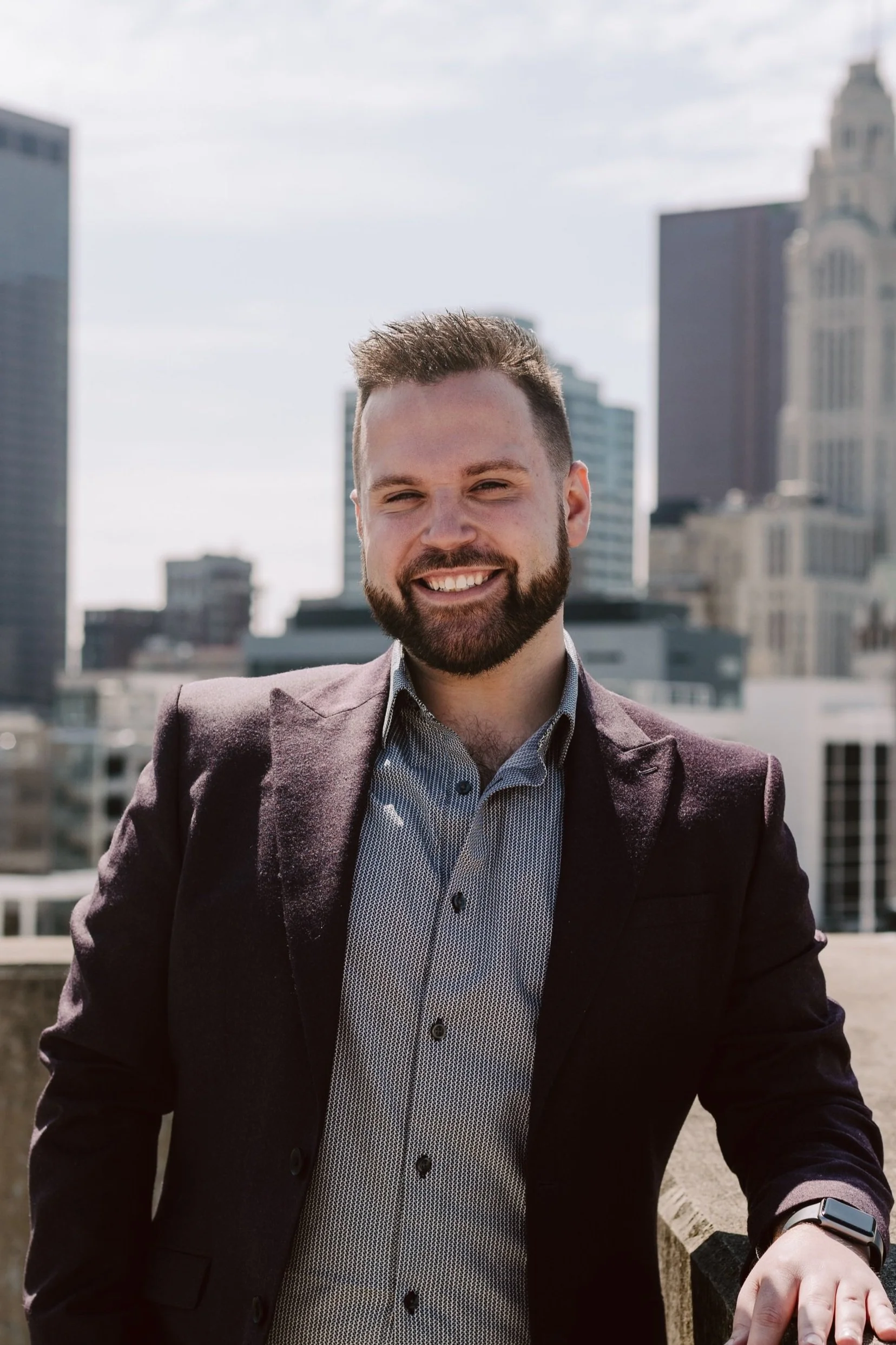 A smiling man with a beard, wearing a dark blazer and light-colored dress shirt, standing on a rooftop in a city with tall buildings in the background.