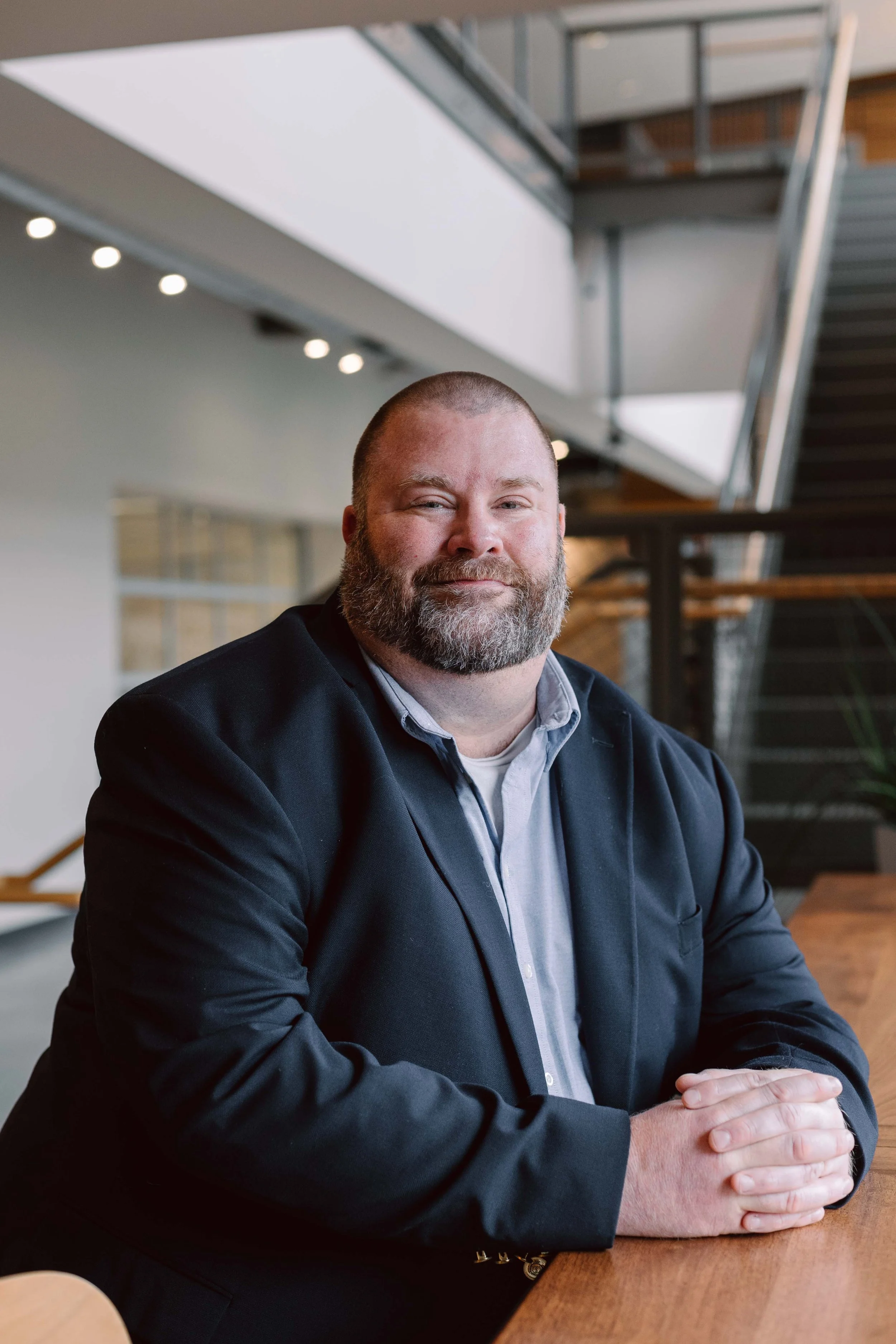 A man with a beard and short hair, wearing a suit jacket and shirt, sitting at a wooden table inside a modern office building with a staircase and railing in the background.
