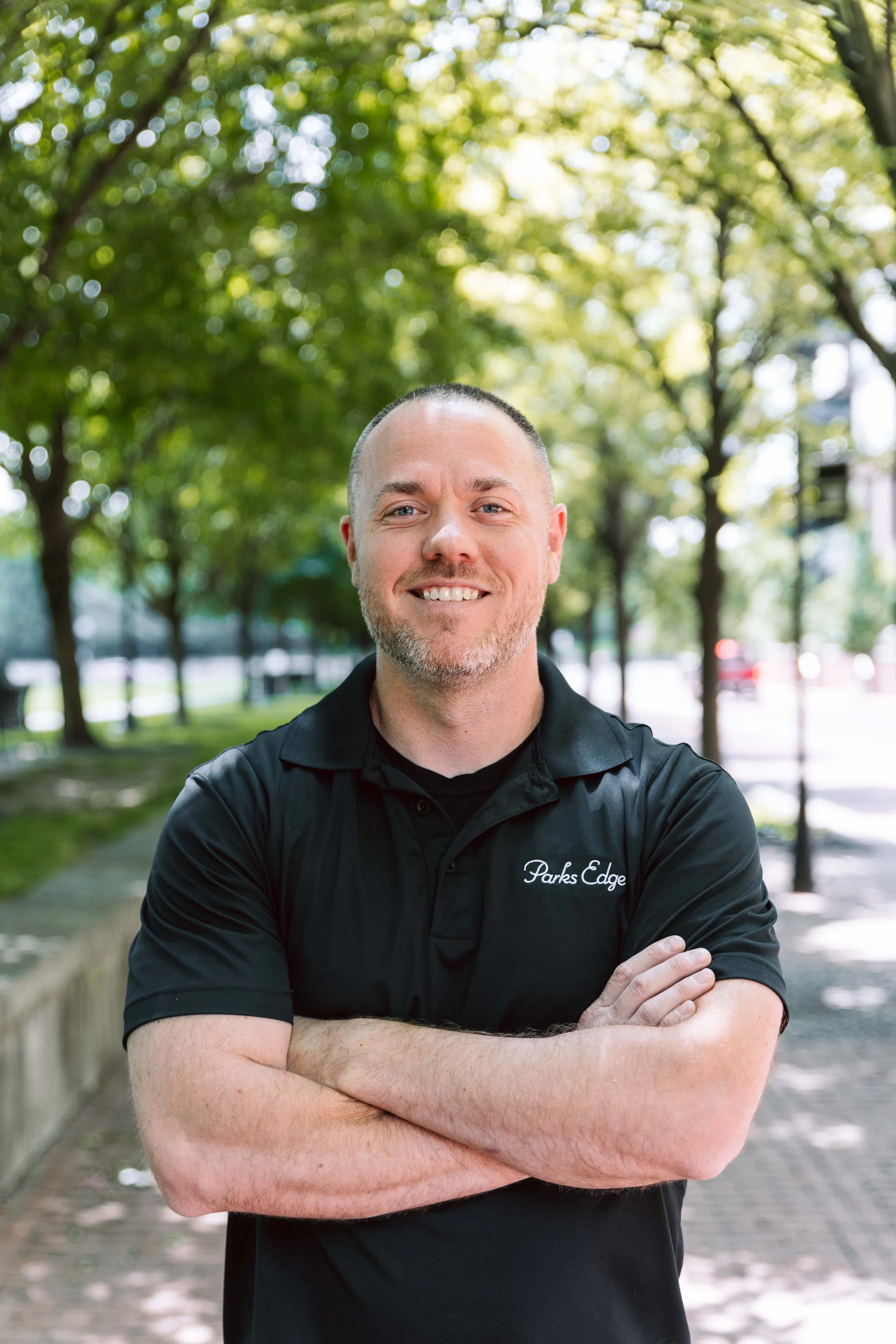 A man with a short beard and a shaved head smiling with arms crossed, standing outdoors in a park with green trees and a sidewalk.