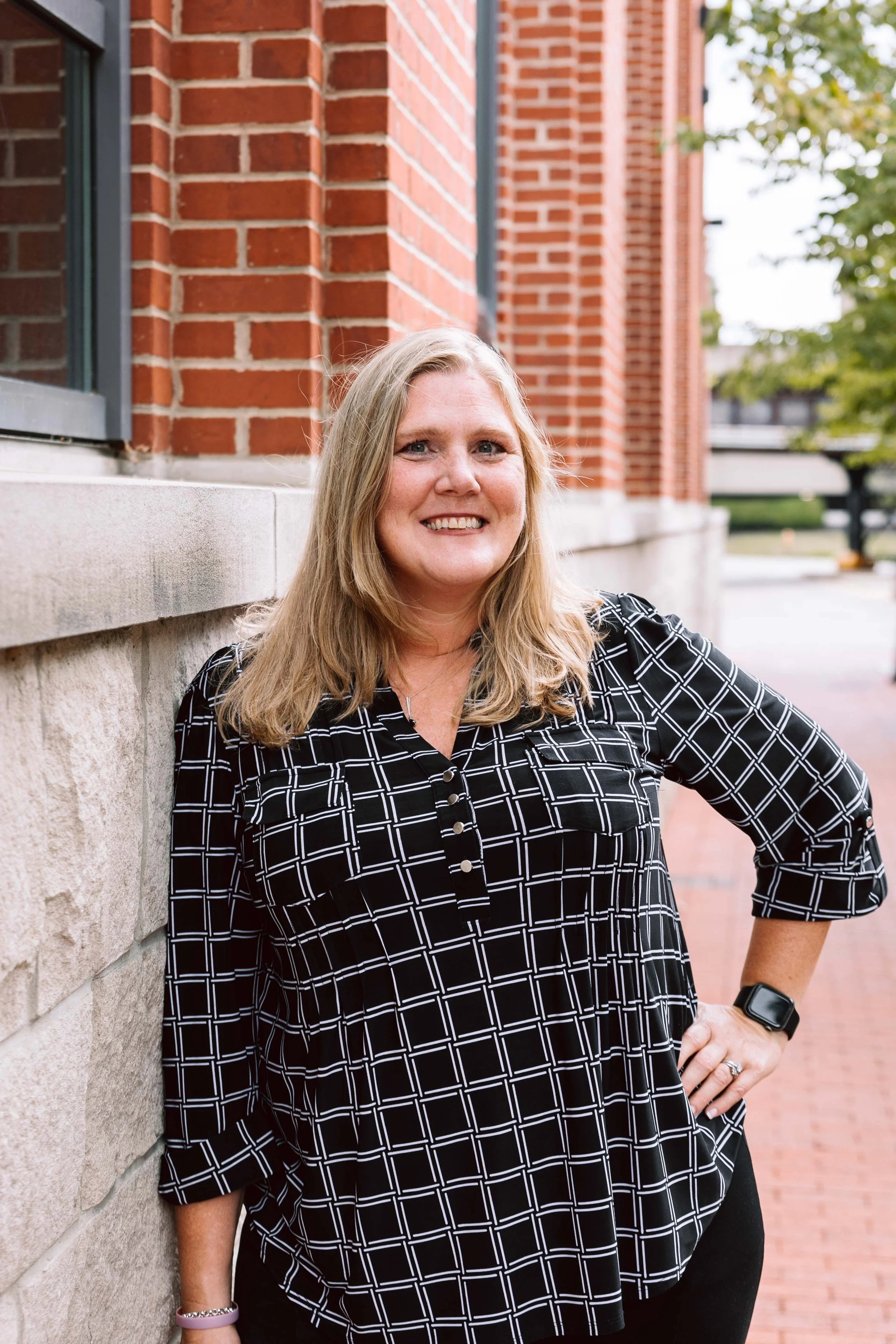 A woman with blonde hair smiling, standing outdoors against a brick wall with her hand on her hip.