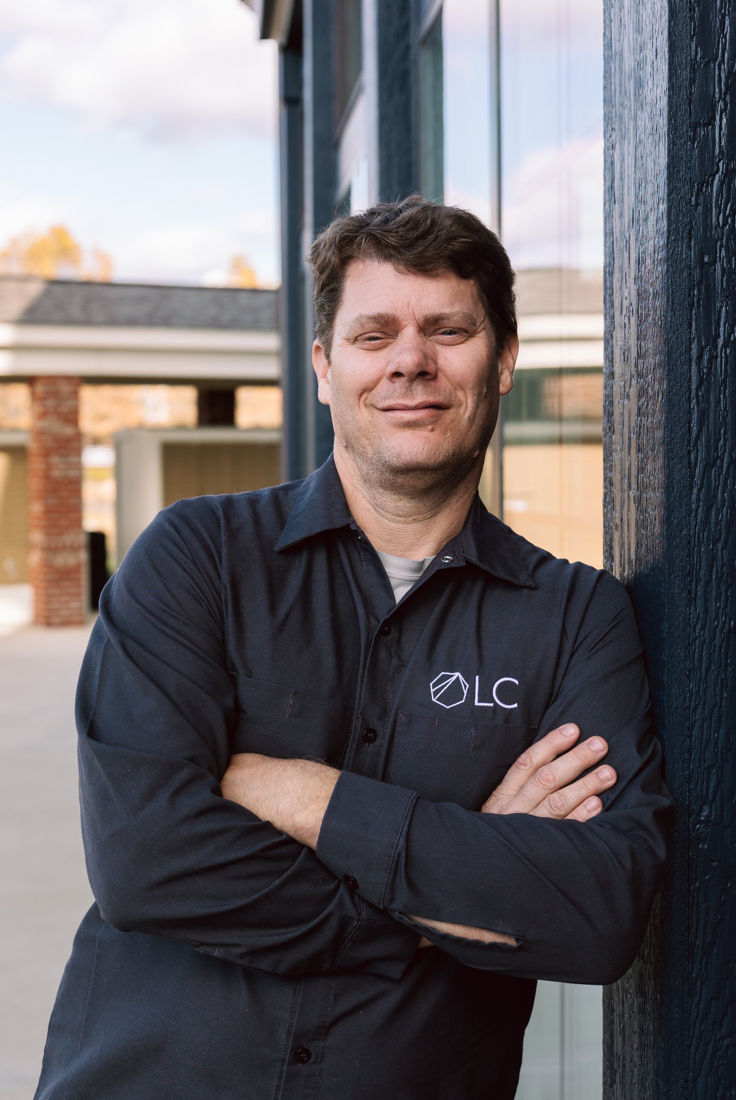 A man with brown hair, smiling, wearing a black shirt with a logo that says 'OLC,' standing outdoors against a building with reflective windows.