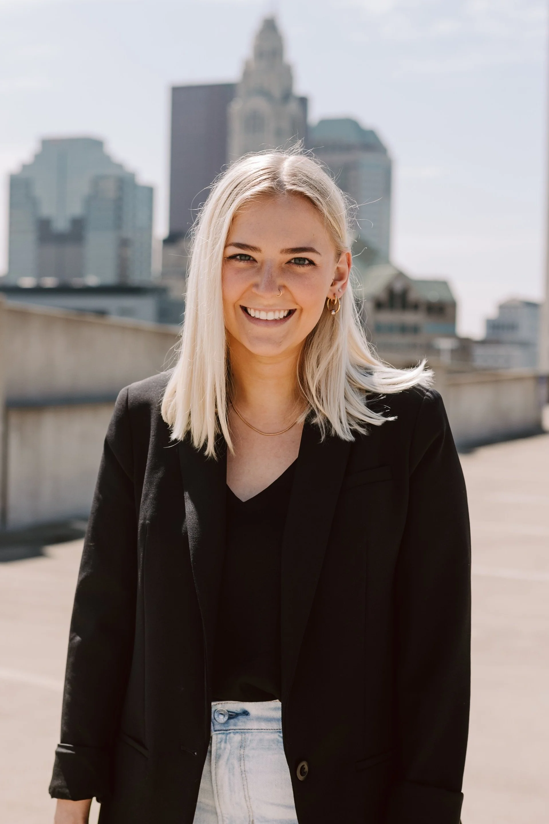A woman with blonde hair smiling outdoors in a cityscape background with tall buildings.