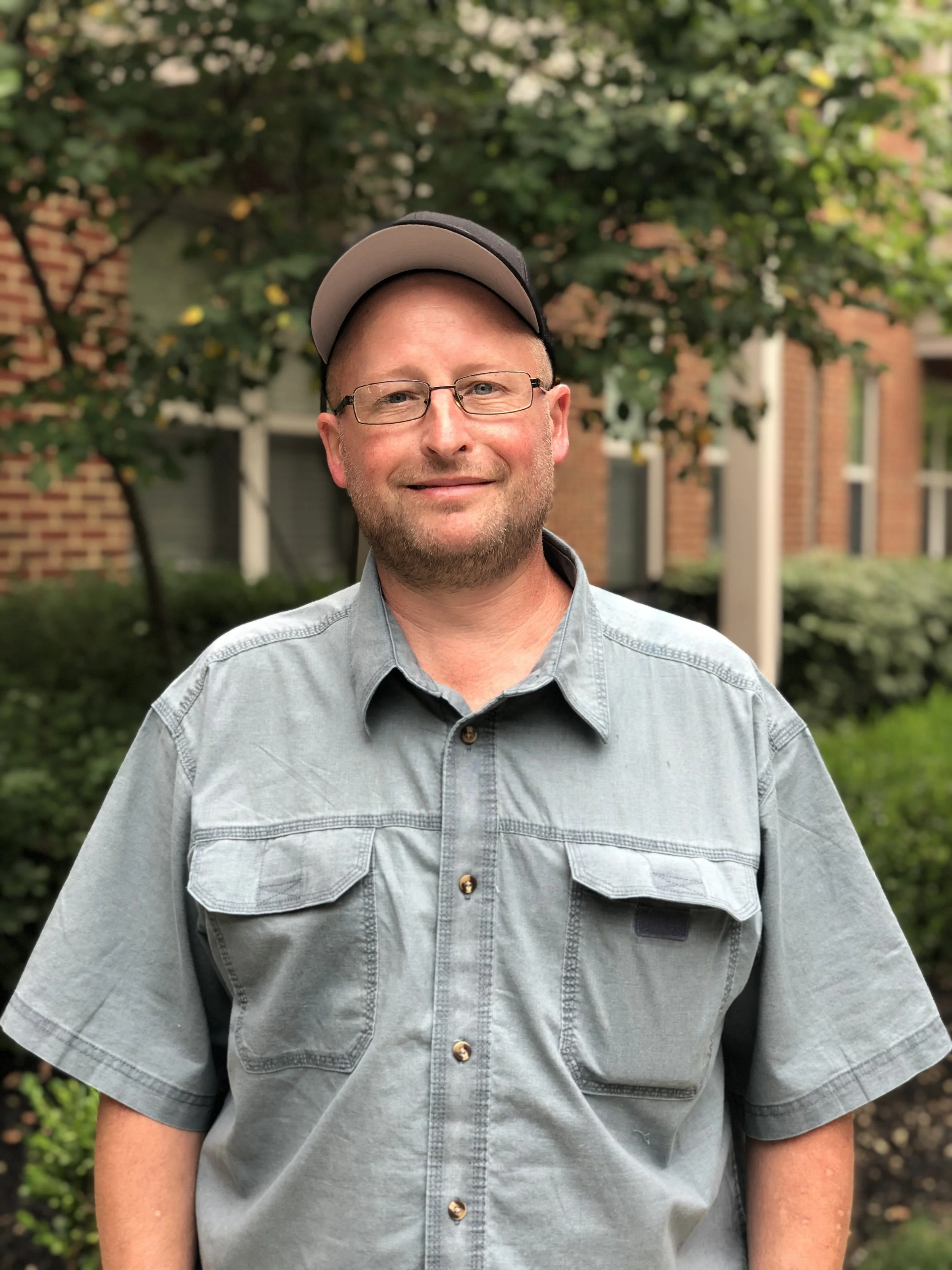 A man with glasses and a beard standing outdoors in front of trees and a brick building, wearing a gray short-sleeved shirt and a gray baseball cap.