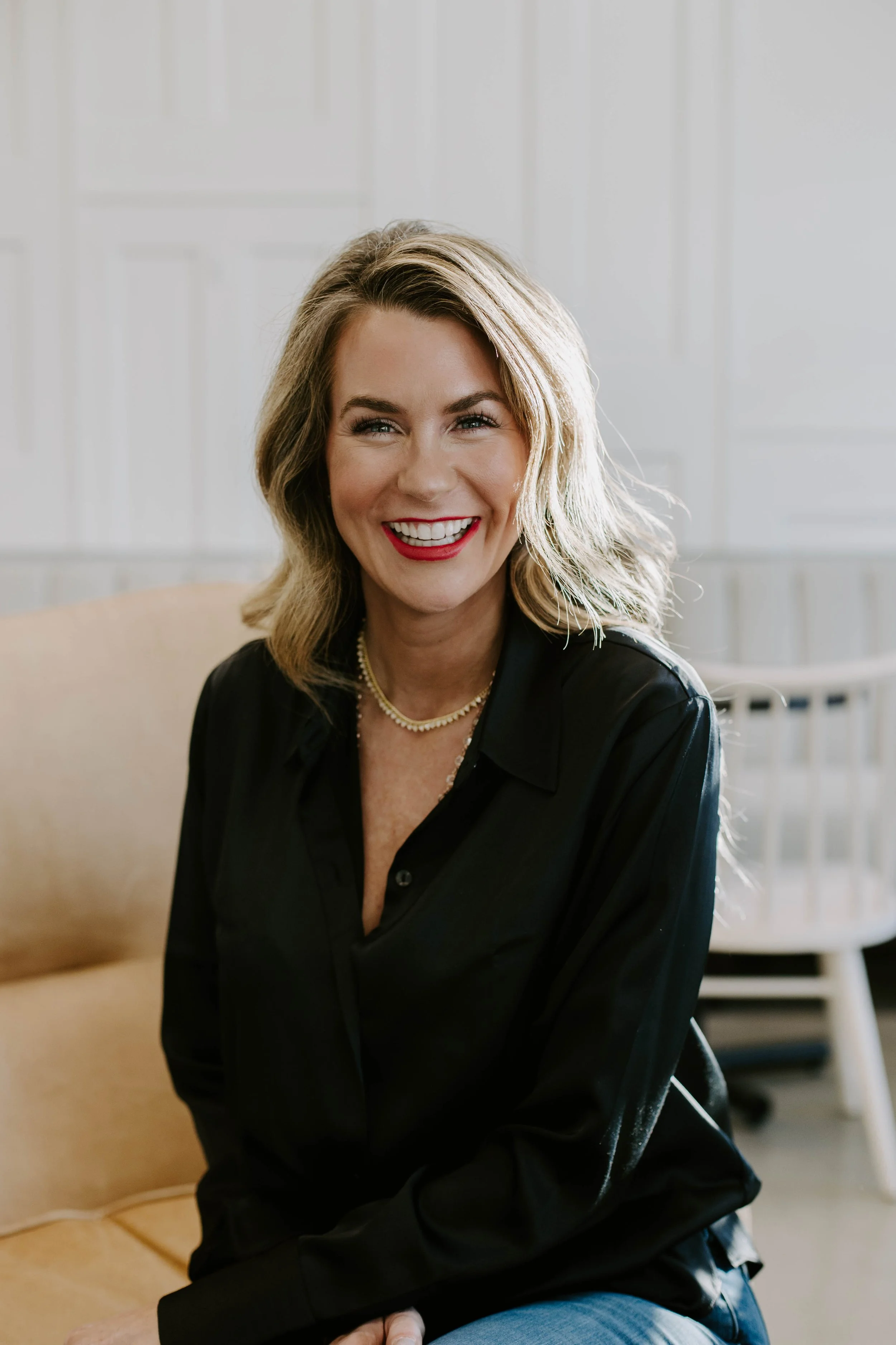A woman with shoulder-length blonde hair, smiling broadly, wearing a black shirt and pearl necklace, sitting on a beige couch in a bright room.