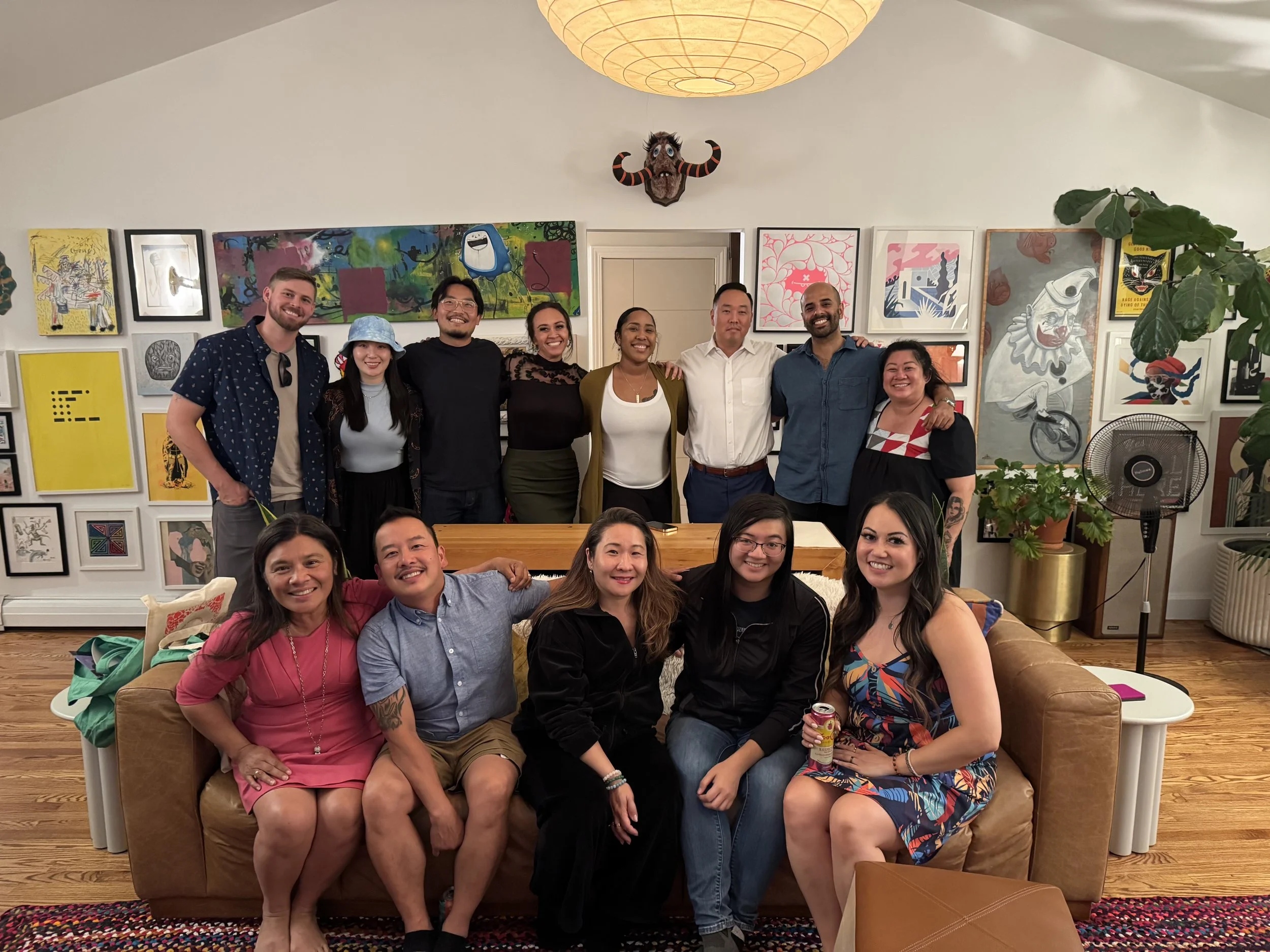 A group of thirteen diverse people smiling and posing in a cozy, art-decorated room. They are seated and standing around a sofa, with a collection of colorful artwork on the walls behind them and a large plant and fan on the side.