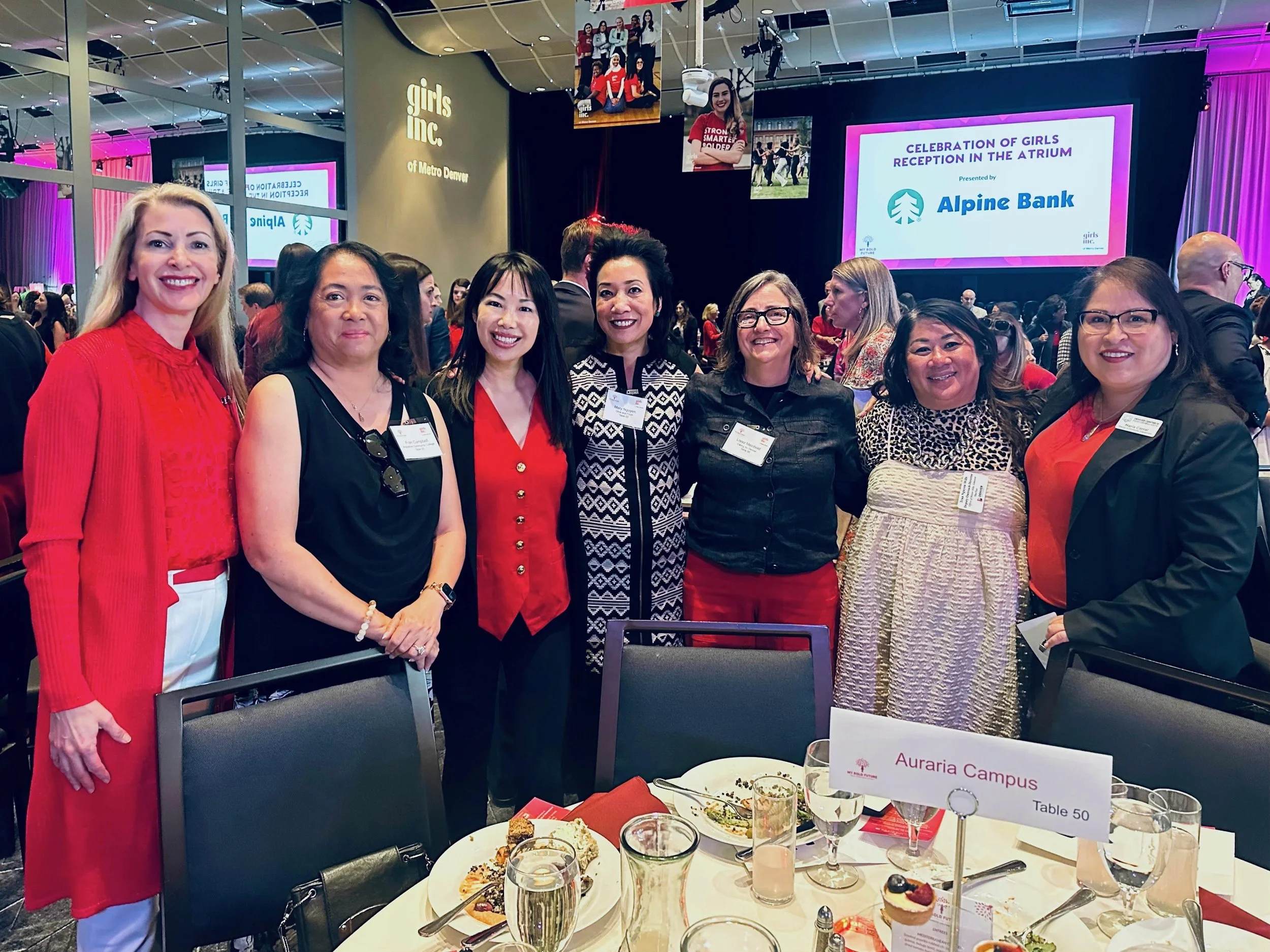 Group of seven women standing together at a conference event, smiling. They are dressed in business casual attire, with some wearing red and black. Behind them, a stage displays the text 'Celebration of Girls Reception in the Atrium' presented by Alp