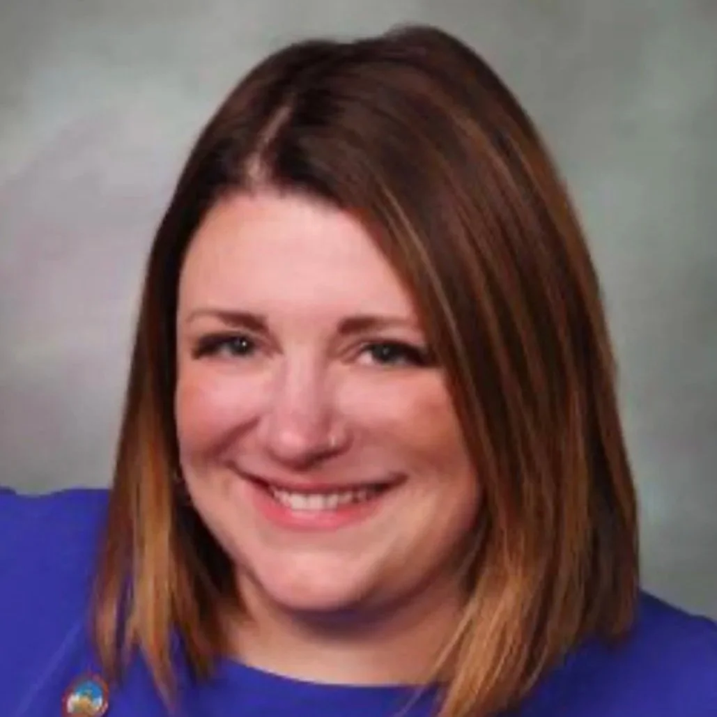 A woman with shoulder-length reddish-brown hair, wearing a blue top, smiling at the camera against a neutral background.