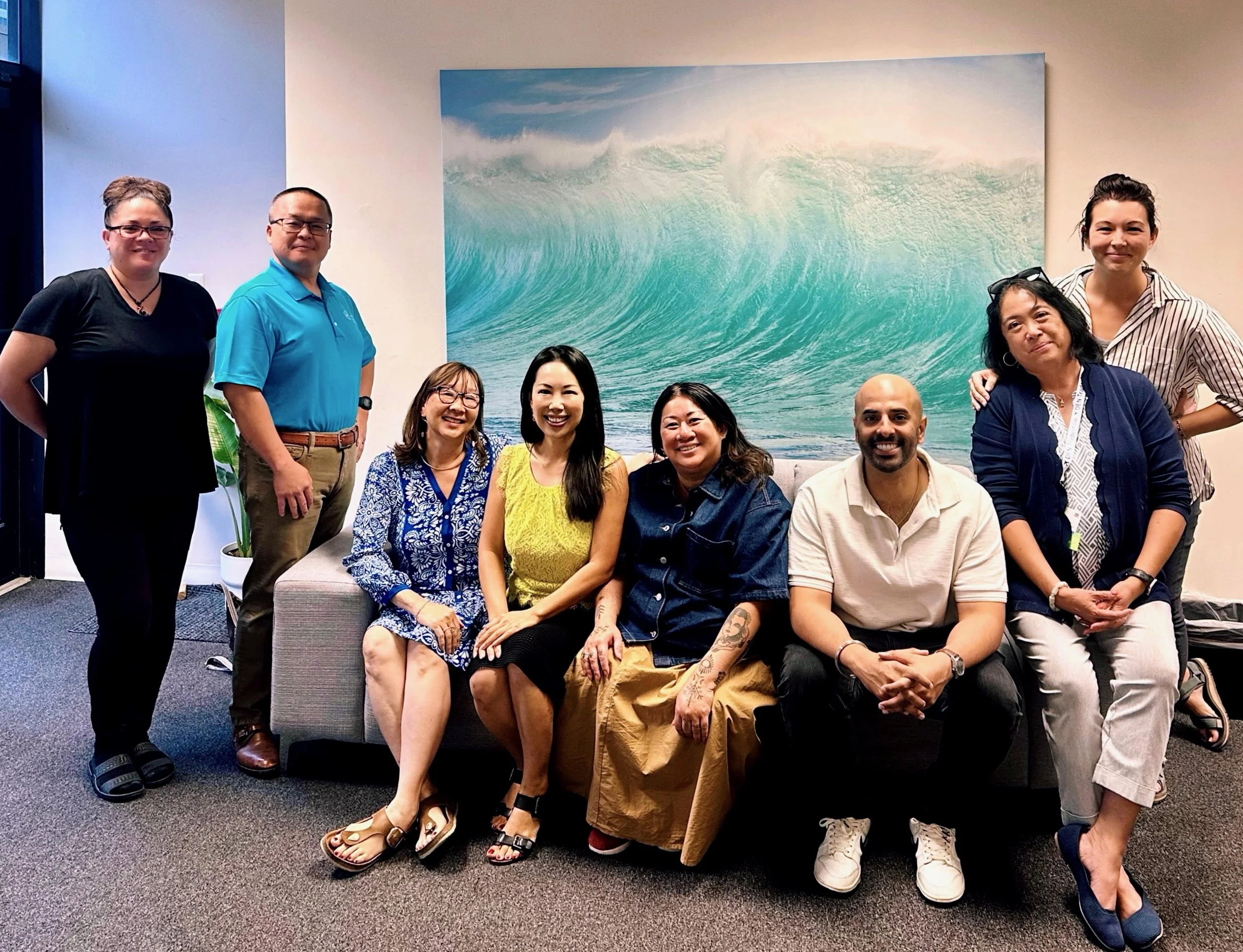 A group of nine diverse people posing in an office, sitting and standing in front of a large ocean wave painting on the wall.