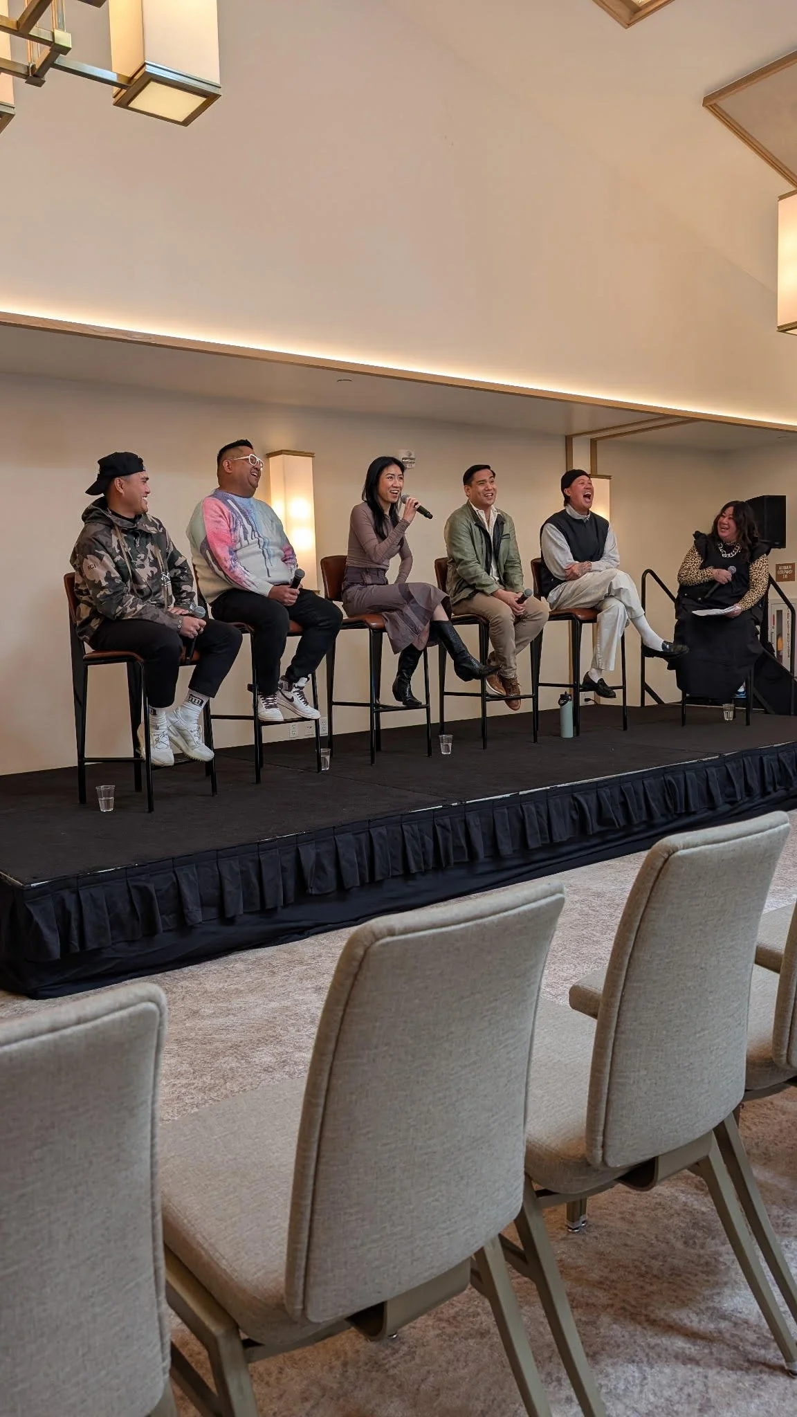 Six people sitting on a stage, engaging in a panel discussion or interview. Five are seated on high chairs, with one person holding a microphone and speaking. The background is a neutral wall with warm lighting, and there are empty chairs in the fore