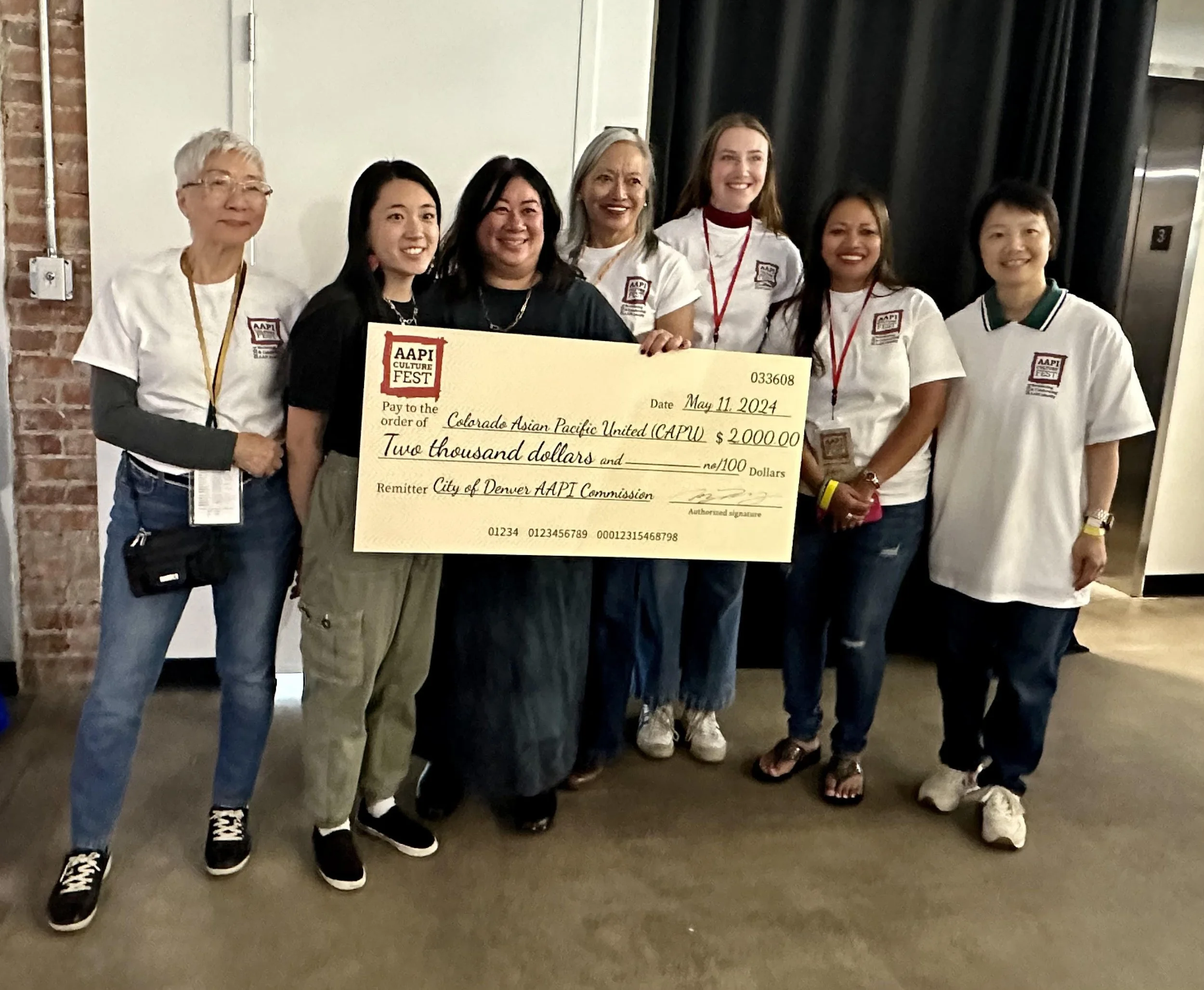Group of seven women holding a large check at an indoor event. The check is made out to Colorado Asian Pacific United for $2,000, dated May 11, 2024. The women are smiling and wearing casual clothing, with some in white t-shirts with a logo.