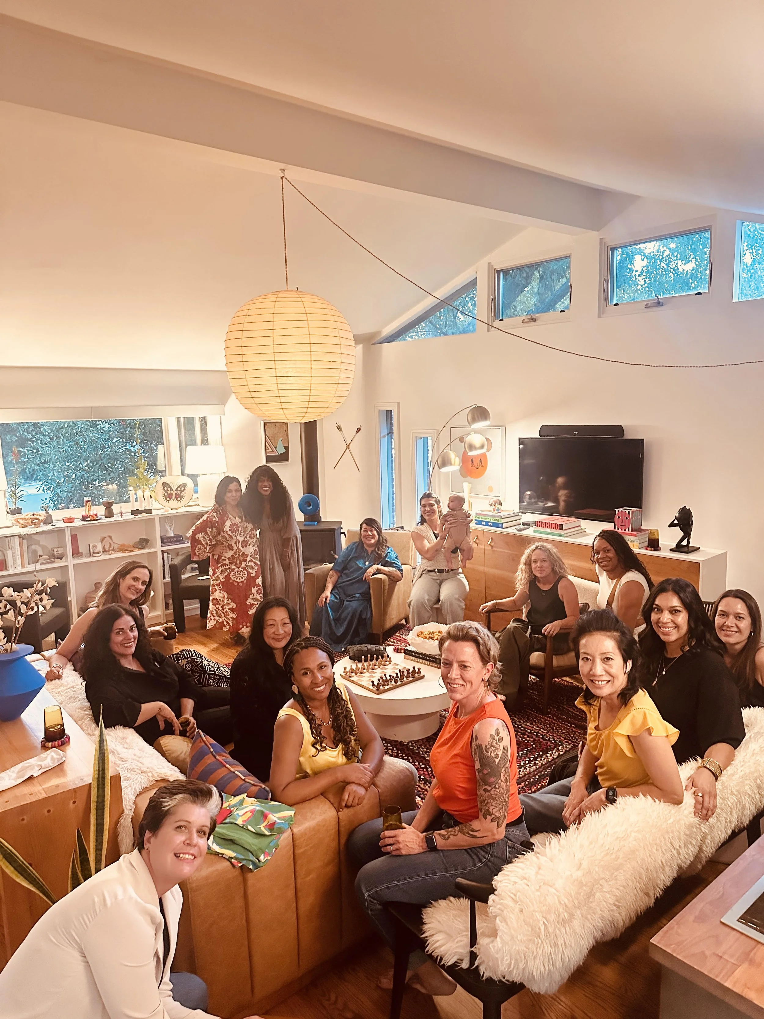Group of women gathered in a modern, well-lit living room, smiling and sitting around a coffee table with a chess set, celebrating a gathering or party.