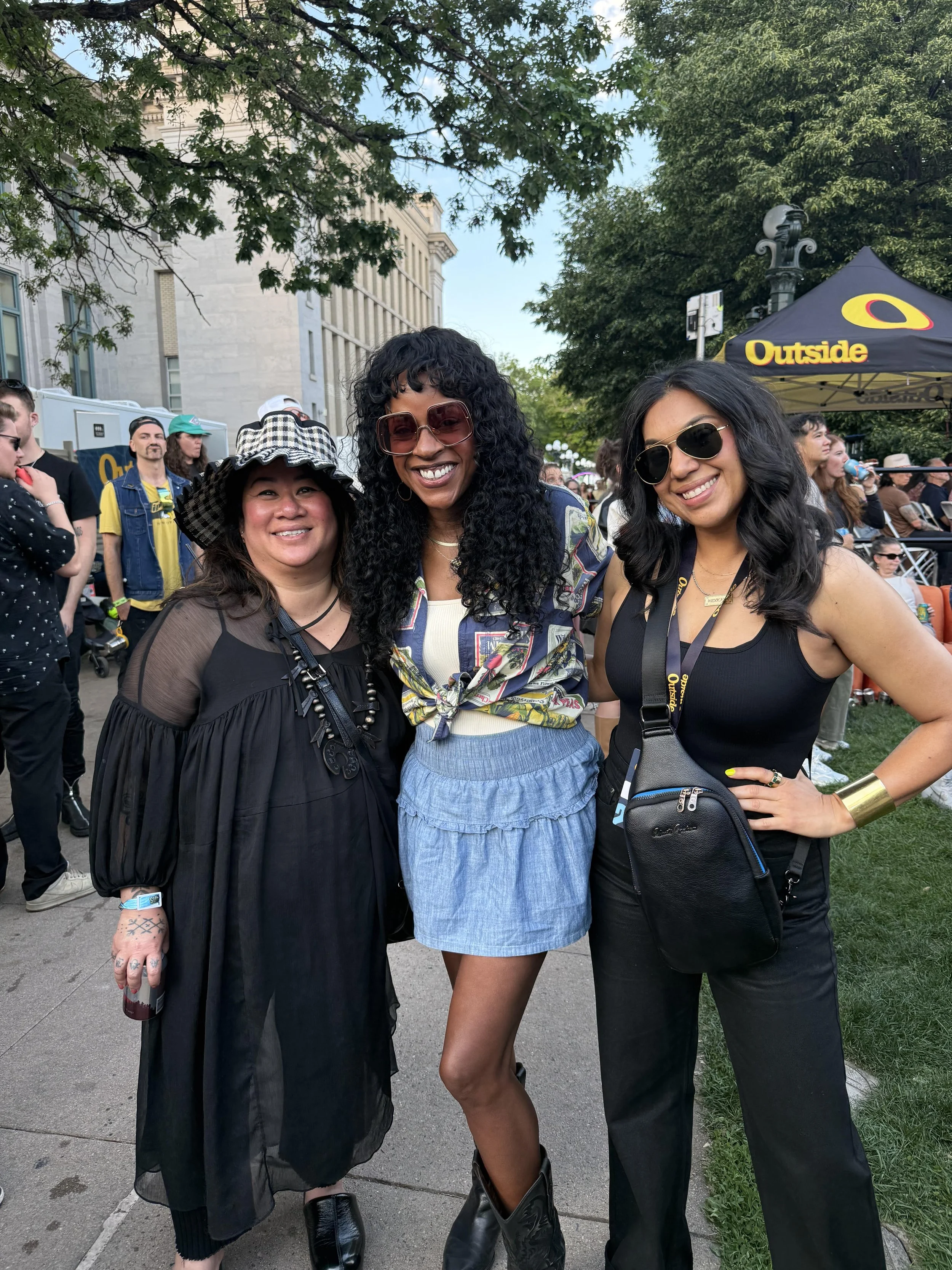 Three women standing together outdoors at an event, smiling at the camera. Two women are wearing sunglasses, and there are trees, a building, and a black tent with yellow lettering in the background.