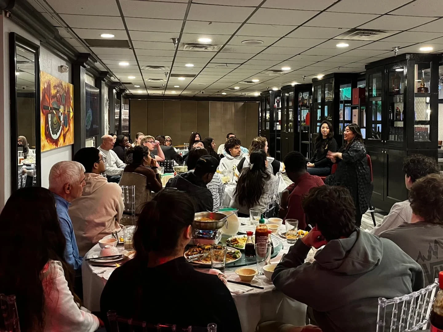 People seated at a dining table listening to two women speaking in a restaurant or banquet hall.
