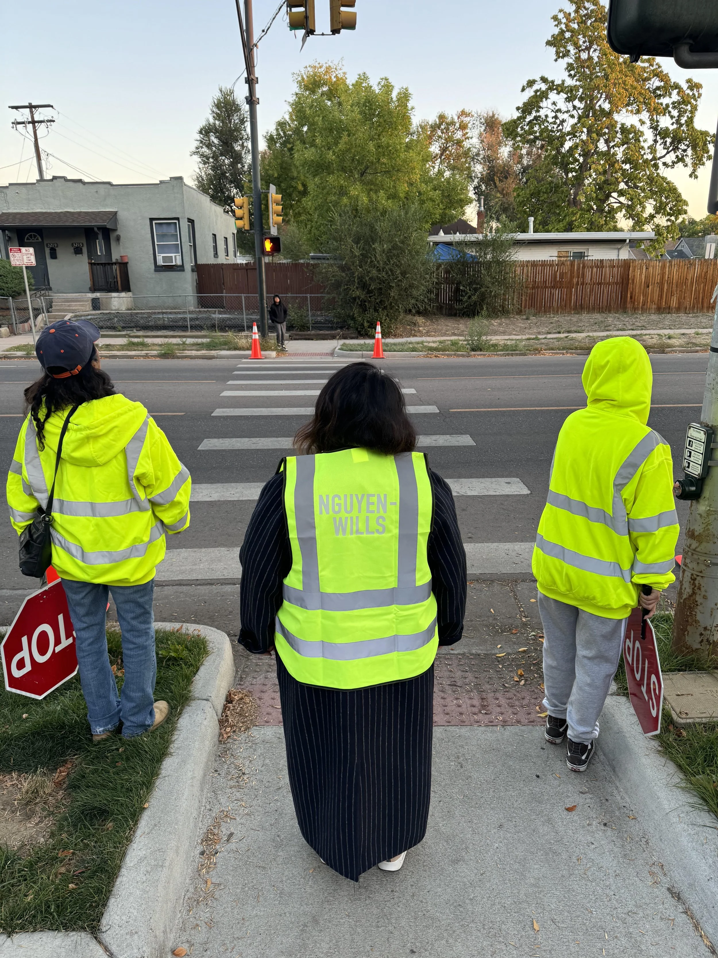 Three people in yellow safety vests holding stop signs at a crosswalk, waiting for traffic to stop at a pedestrian crossing.