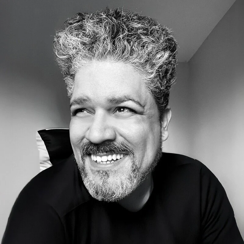 Black and white photo of a smiling man with curly gray hair and a gray beard, wearing a black shirt, sitting in front of a plain wall.
