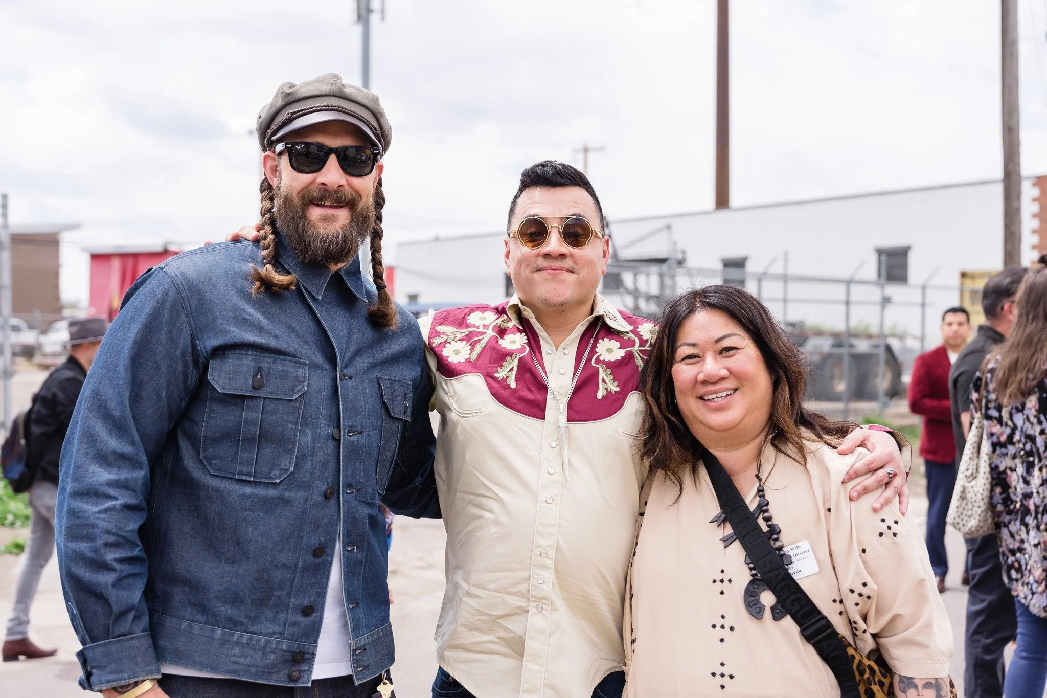 Three diverse people smiling and posing together outdoors, with a group of people and industrial buildings in the background.