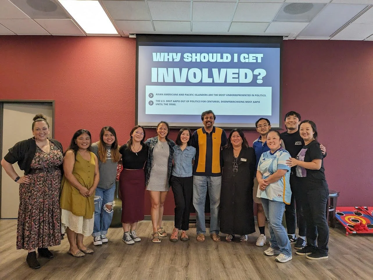Group of people standing in front of a presentation screen that reads "Why Should I Get Involved?" in a room with maroon walls.