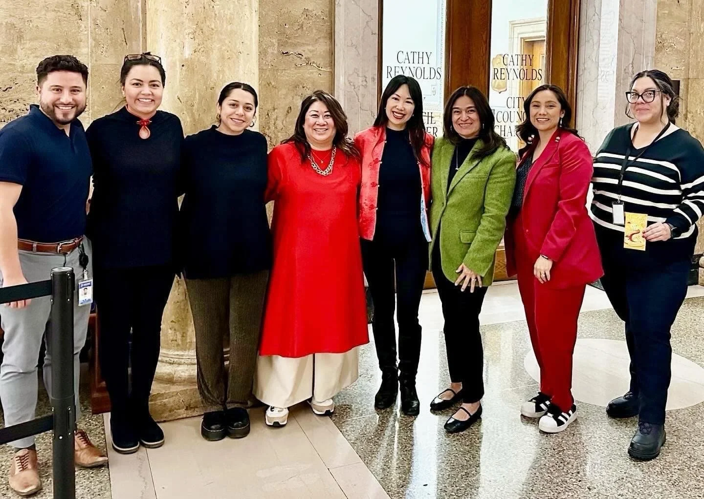 Group of nine people standing together indoors, smiling, in front of a marble wall and wooden door with signs that read 'Cathy Reynolds' and 'City Council'.