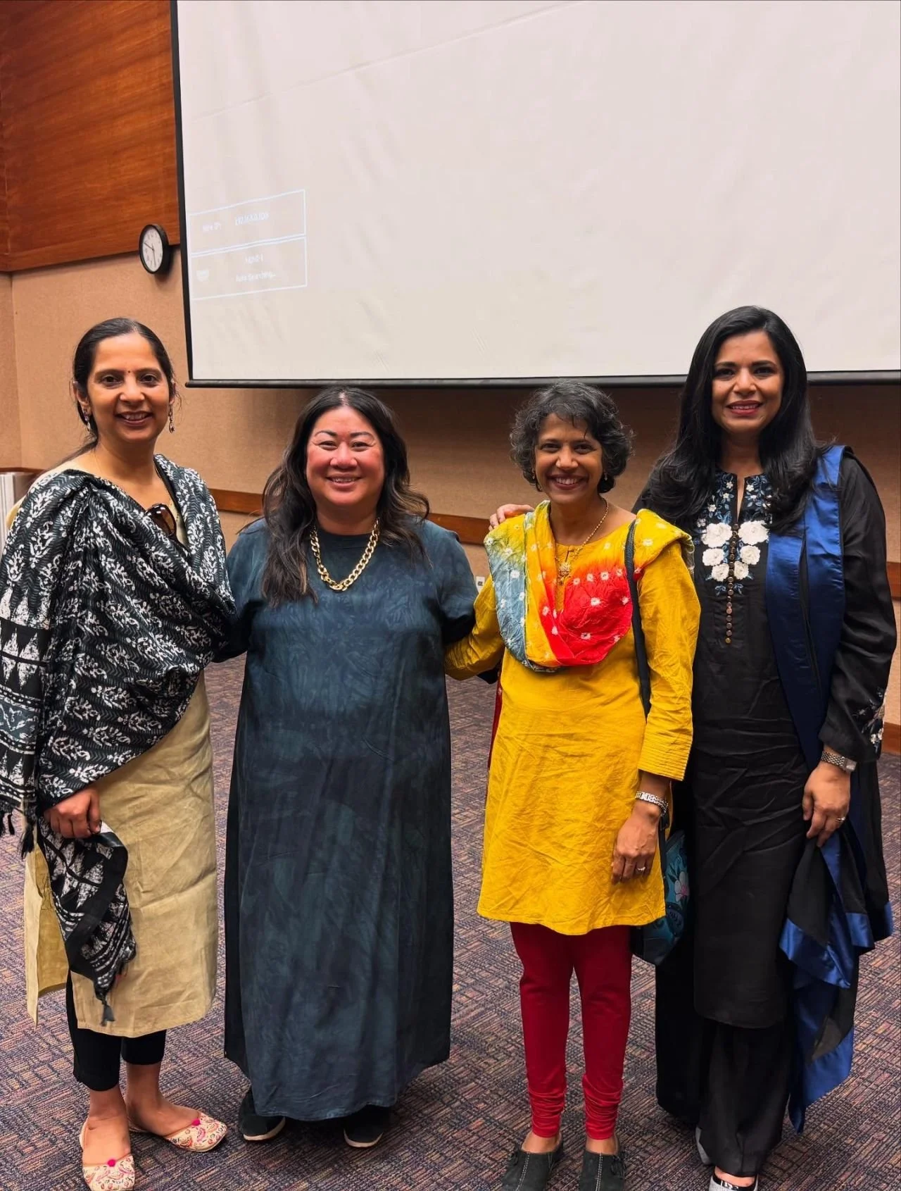 Four women standing together in front of a large presentation screen in a conference room, smiling at the camera.