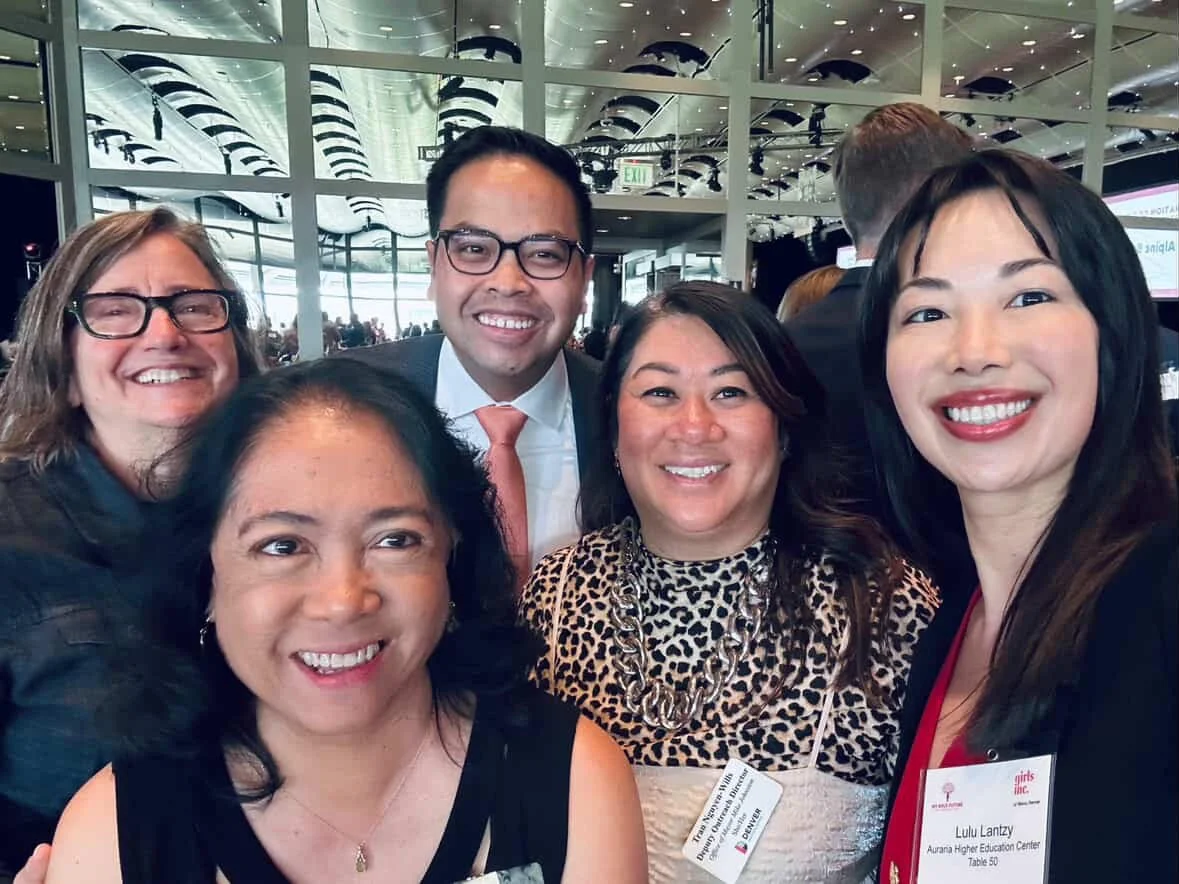 A group of five diverse people smiling together at an indoor event, with a modern architectural background and reflective ceiling.