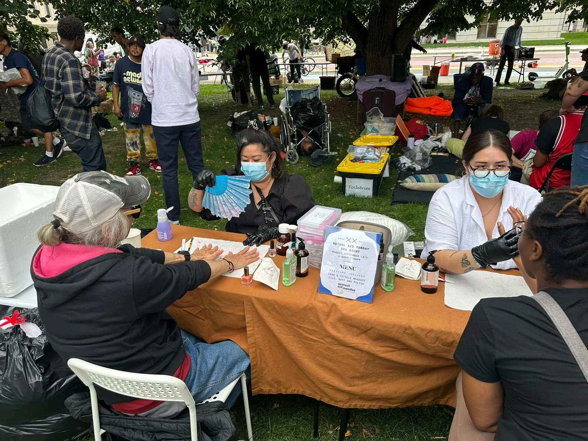 A community event in a park where a woman on the left is getting her nails done by a technician with long black hair, wearing a face mask and gloves. The technician is holding a fan and various nail supplies are on the table. Multiple people are in t